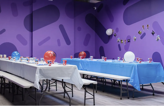 Two rows of party tables with white and blue tablecloths and balloons against a purple wall with a pattern.