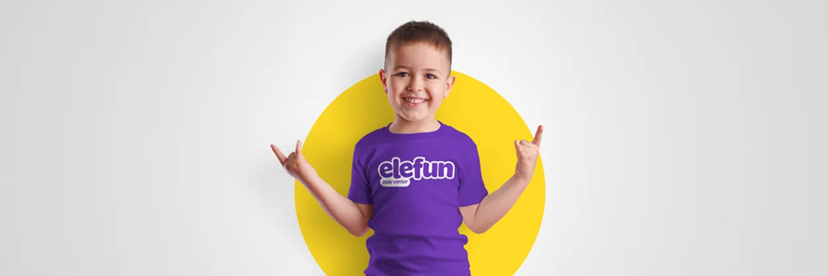 Smiling child in a purple t-shirt with a white graphic standing in front of a yellow circle, making a rock-on hand gesture.