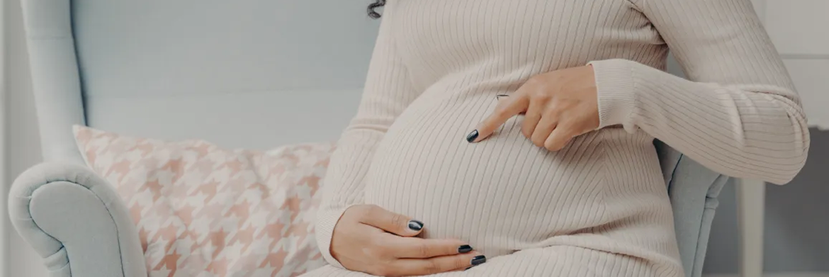 A person wearing a beige long-sleeved dress sits in a light blue chair, gently pointing to their pregnant belly.