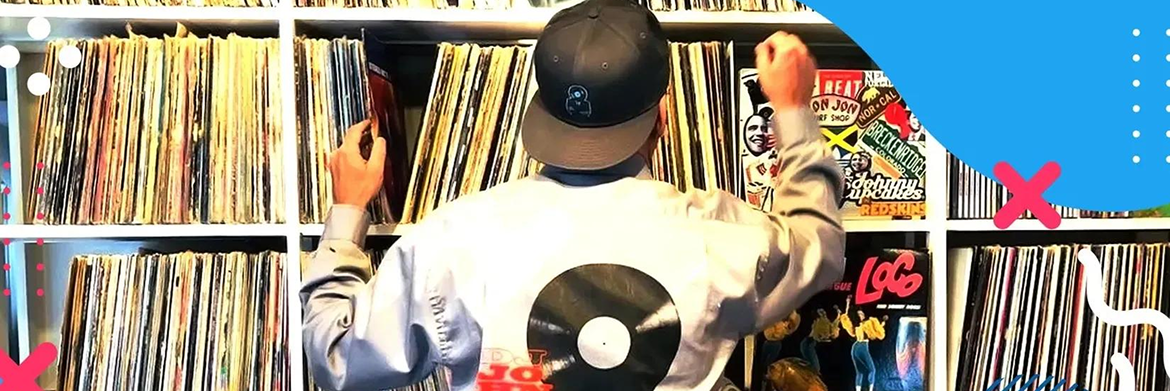 A person wearing a cap stands before floor-to-ceiling shelves filled with vinyl records, reaching to select a disc.