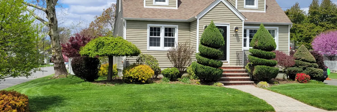 A tan house with a manicured lawn, shaped evergreen shrubs, and flowering trees under a blue sky.