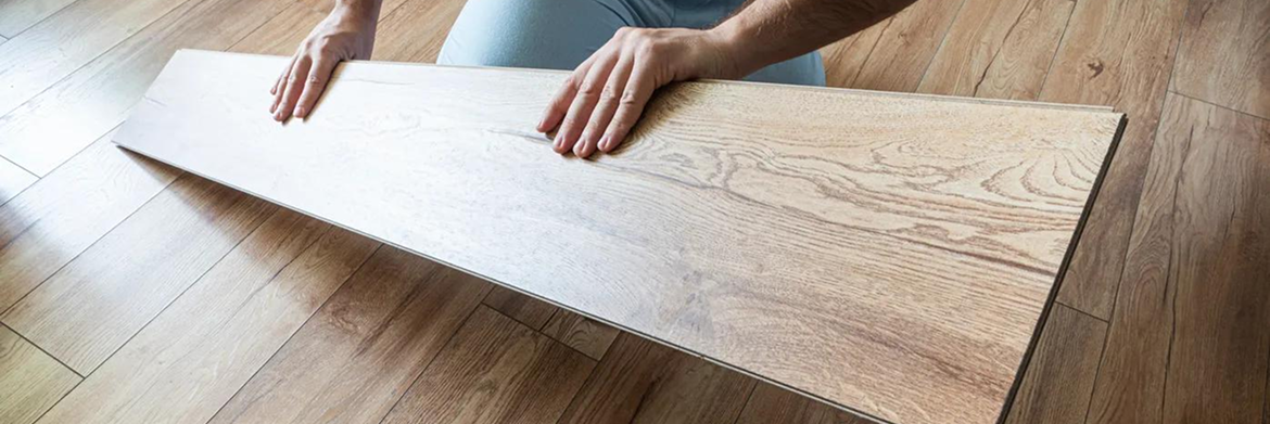 Hands holding a light-colored wood flooring plank against an existing wood floor.