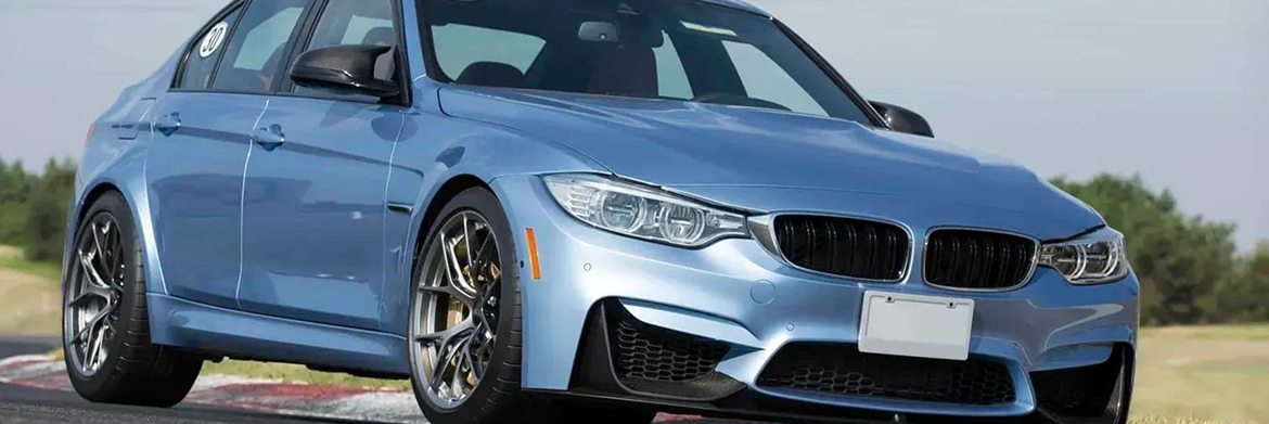 A light blue BMW M3 sedan drives along an asphalt track, showcasing its sporty design and wheels.