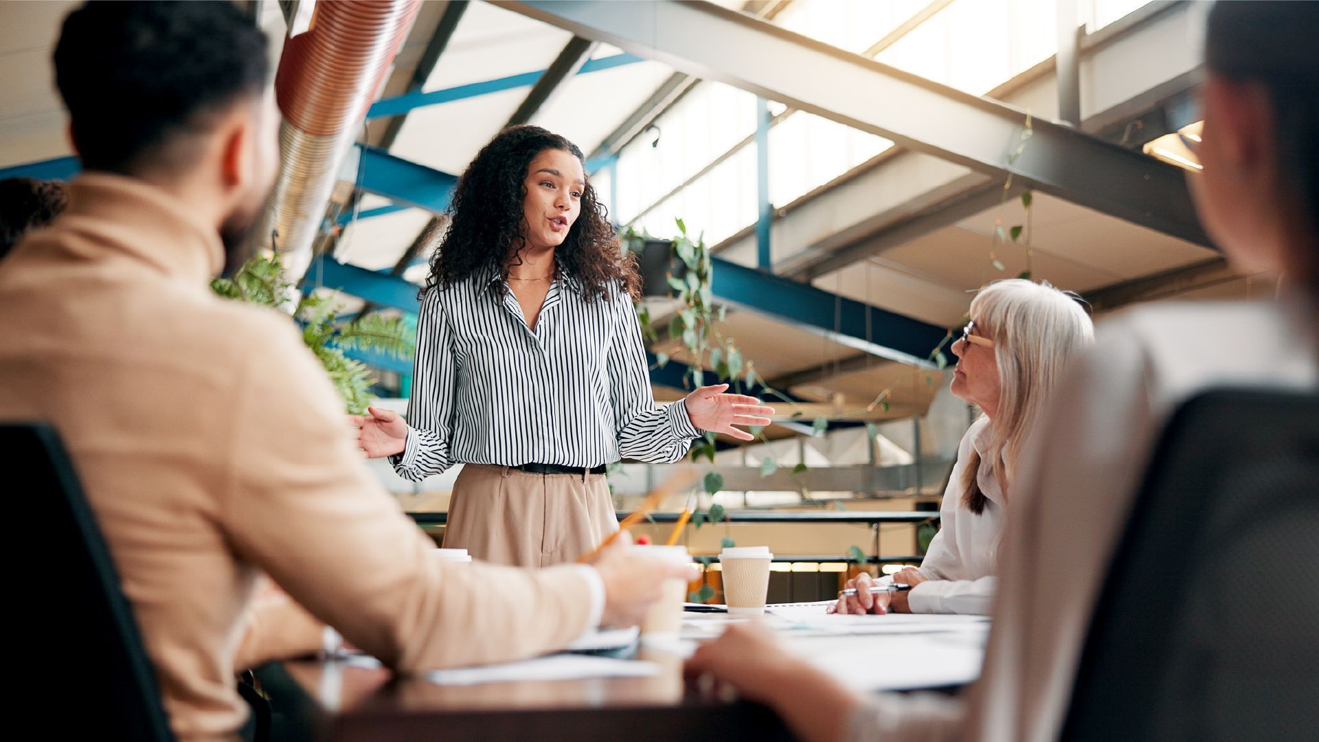 A person in a patterned shirt presents to colleagues seated around a table in a modern, open-plan office.