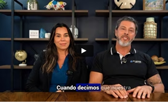 A woman and a man sit at a wooden desk in front of shelves, with Spanish text overlaid at the bottom of the video frame.