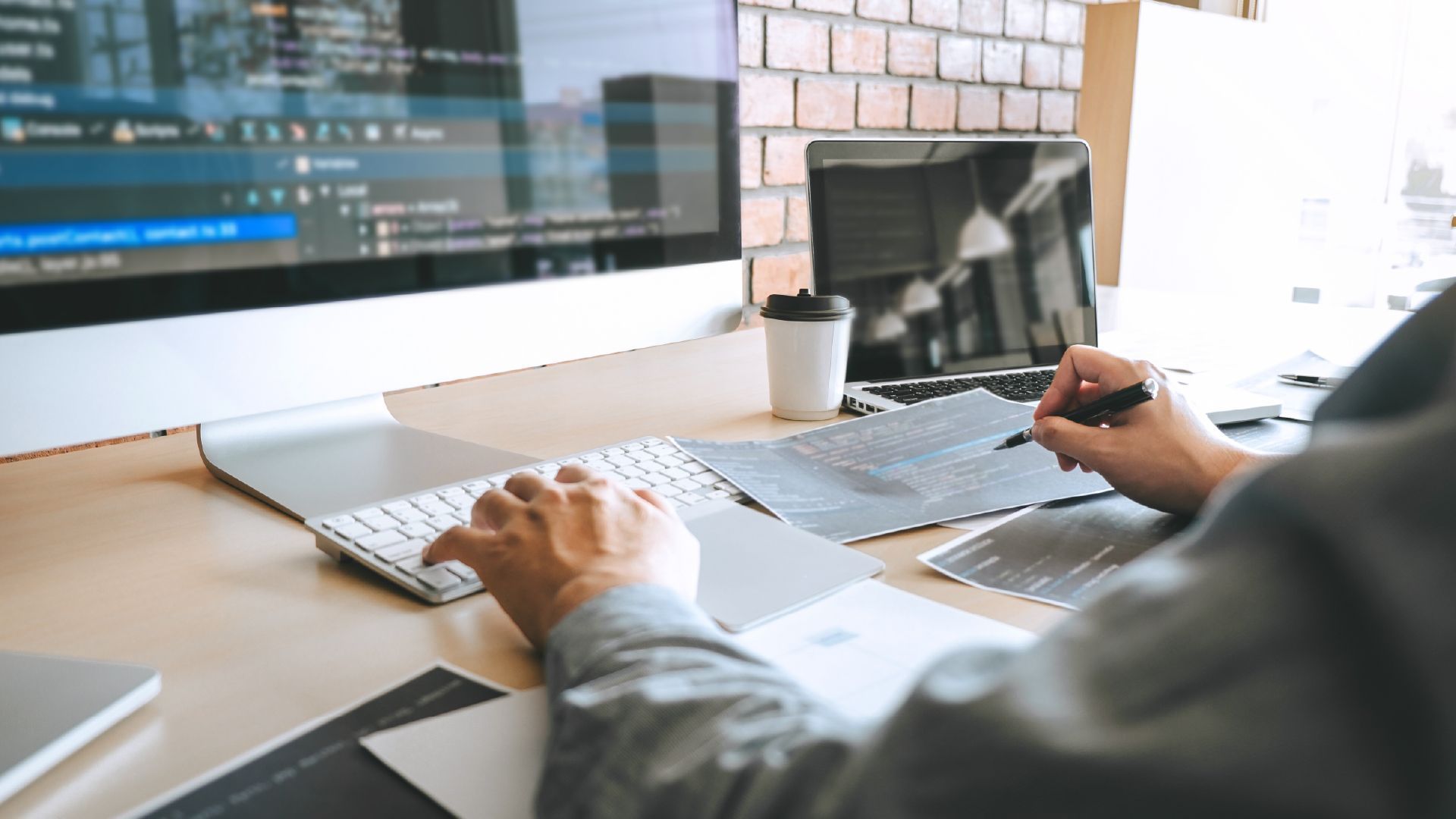 A person typing on a keyboard and taking notes on a notepad at a desk with a computer monitor displaying code.
