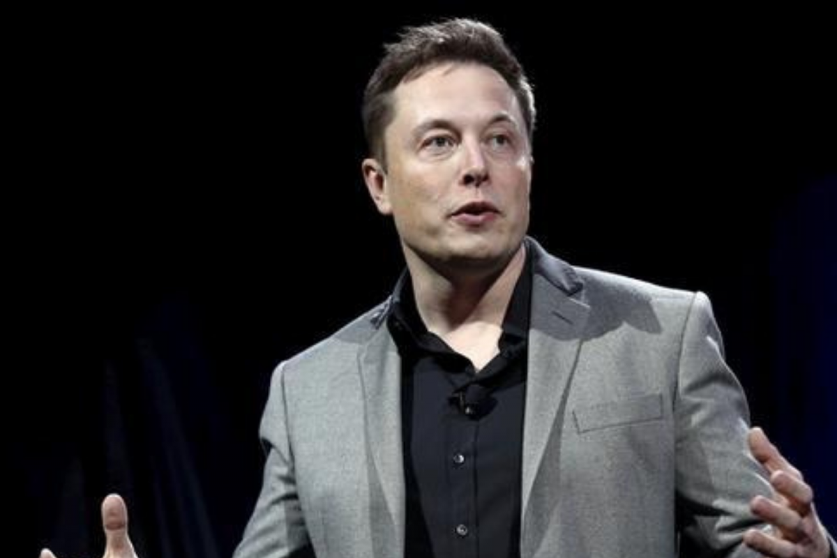 Elon Musk gesturing while speaking against a dark background, wearing a gray blazer over a black button-down shirt.