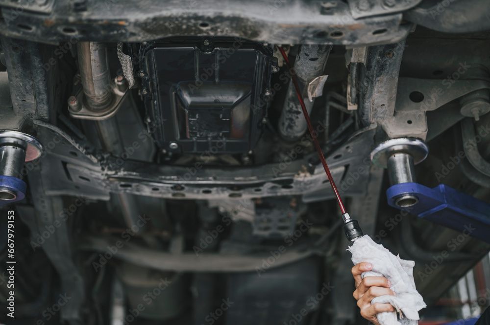 Underside view of a car being serviced. Technician checks fluid levels with a dipstick.