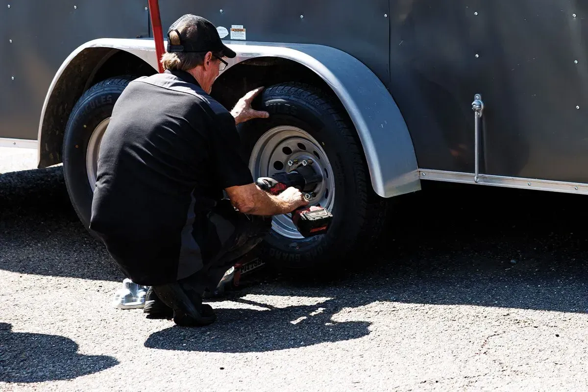 Man in black outfit using a power drill on a trailer tire in an outdoor setting.