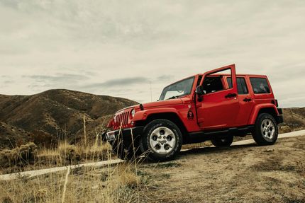 Red Jeep Wrangler parked on a dirt road overlooking rolling hills; door open.