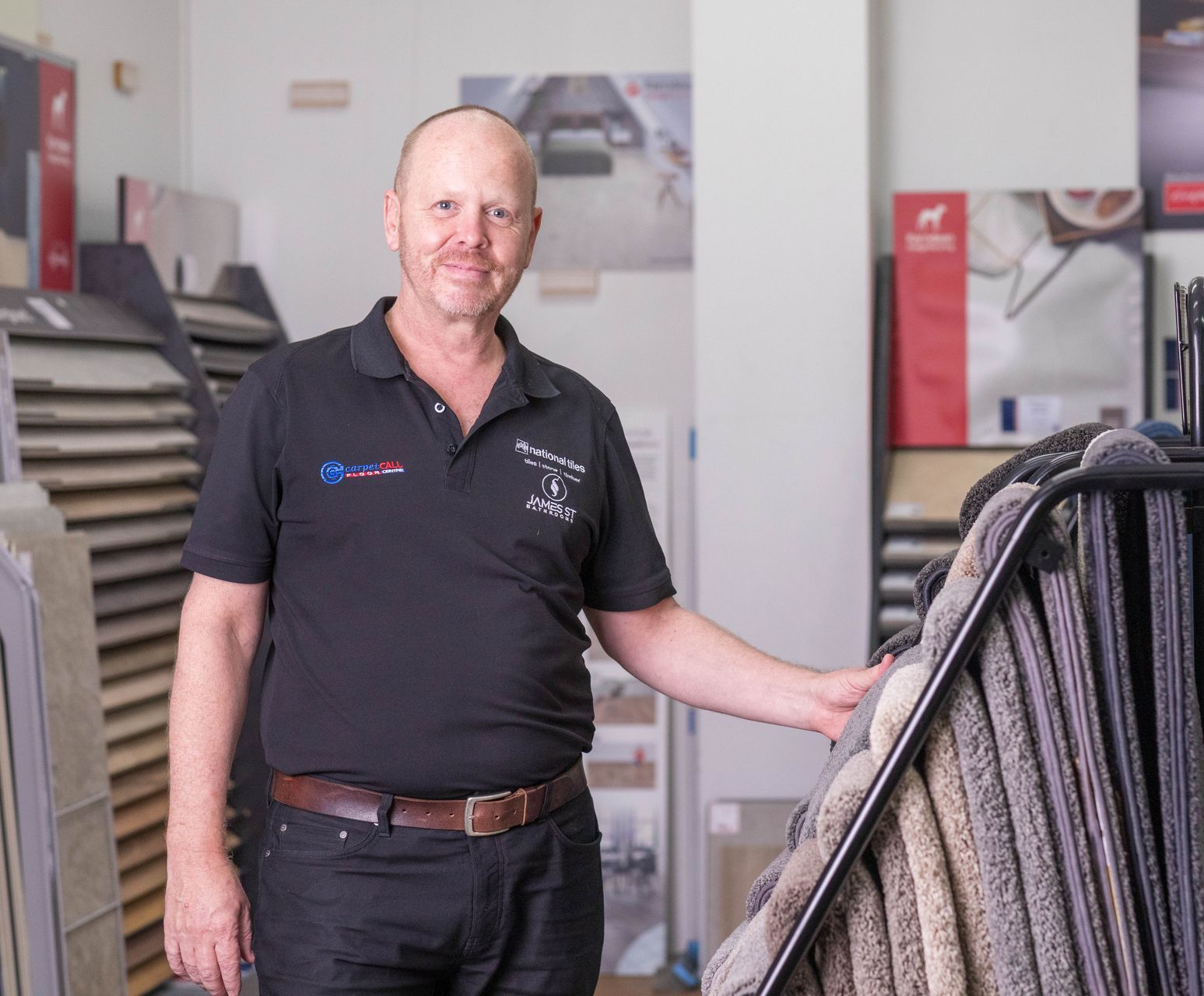 Man in black shirt smiles, standing in a flooring store. He touches a carpet sample rack.