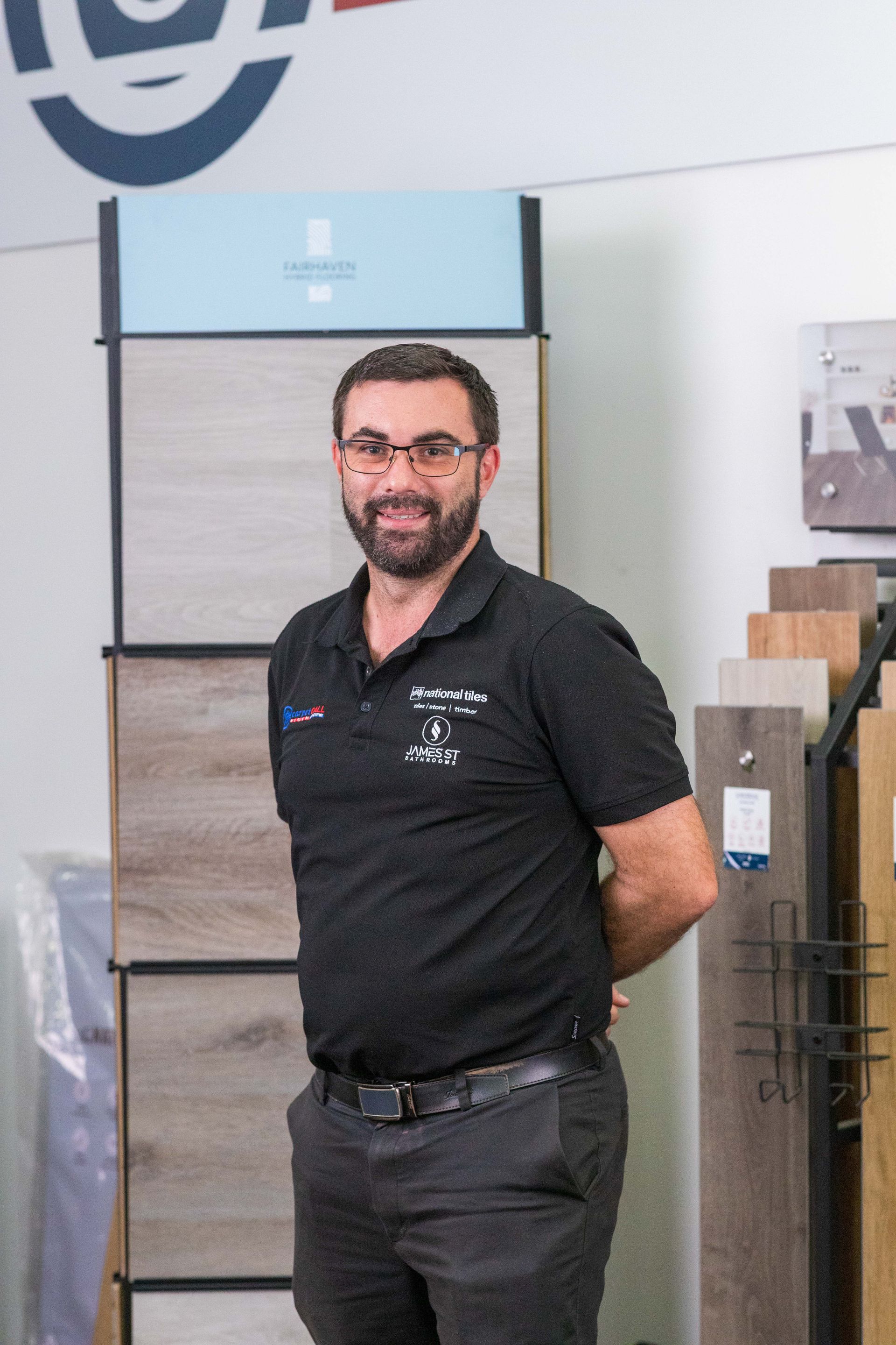 Man with glasses, hands behind back, in a showroom, surrounded by flooring samples.