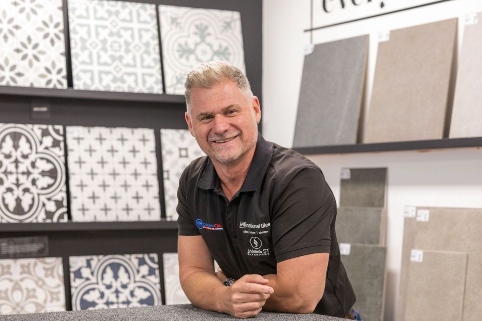 Man in a tile store, smiling, leaning on a counter, with tile samples on the wall.
