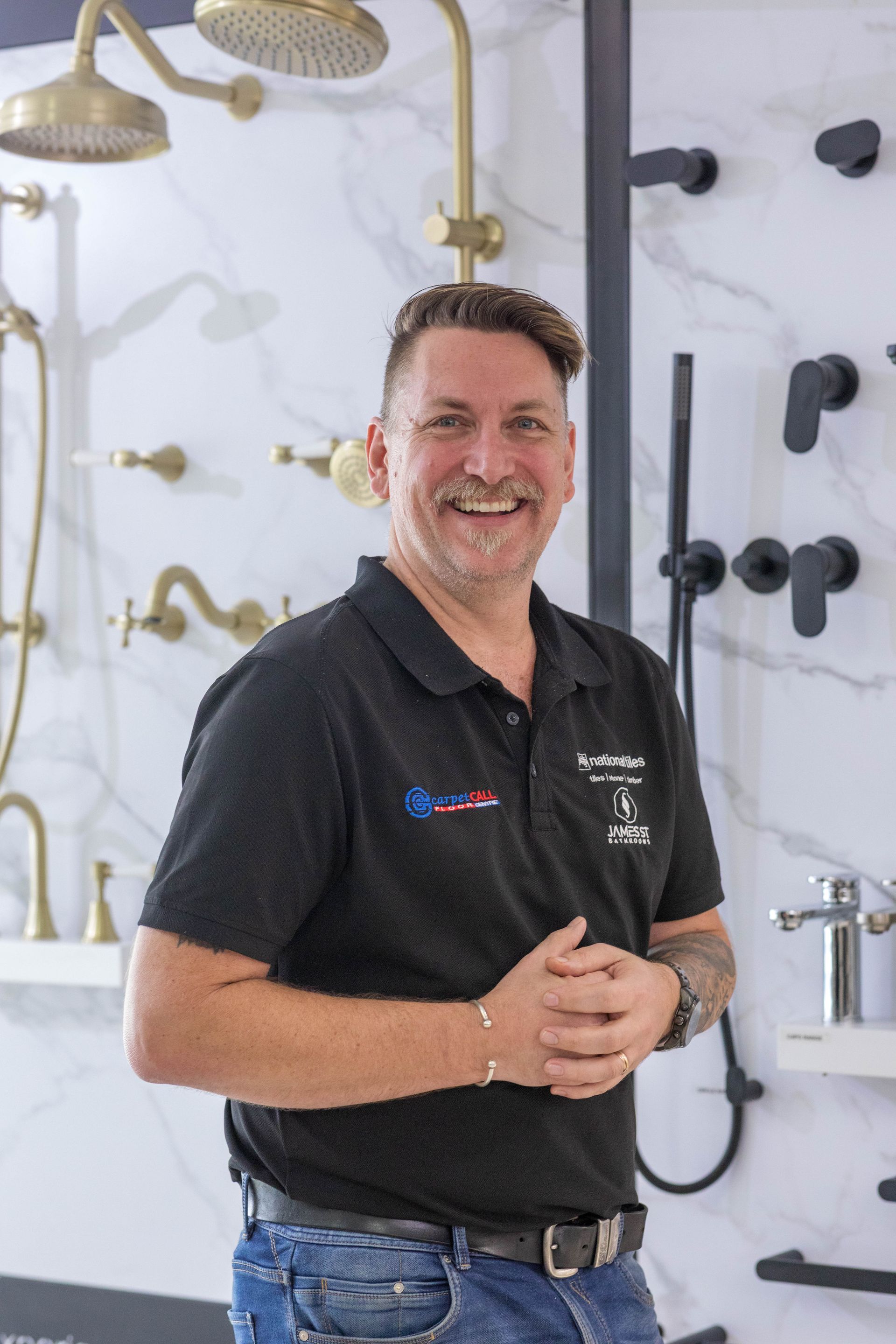 Man smiling in a showroom with bathroom fixtures. He wears a black polo shirt and jeans.