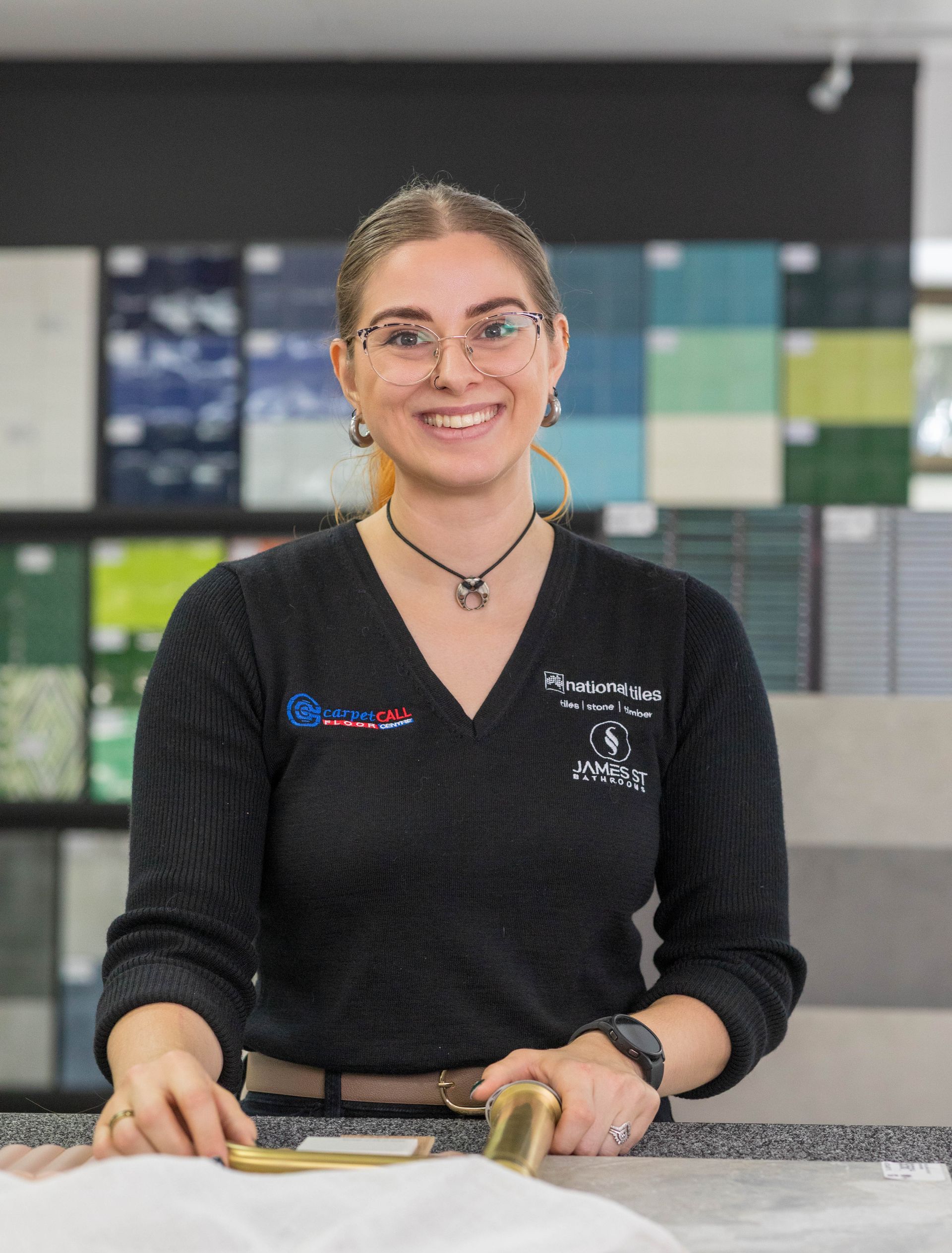 Woman in glasses smiles, holding gold hardware in a tile showroom.