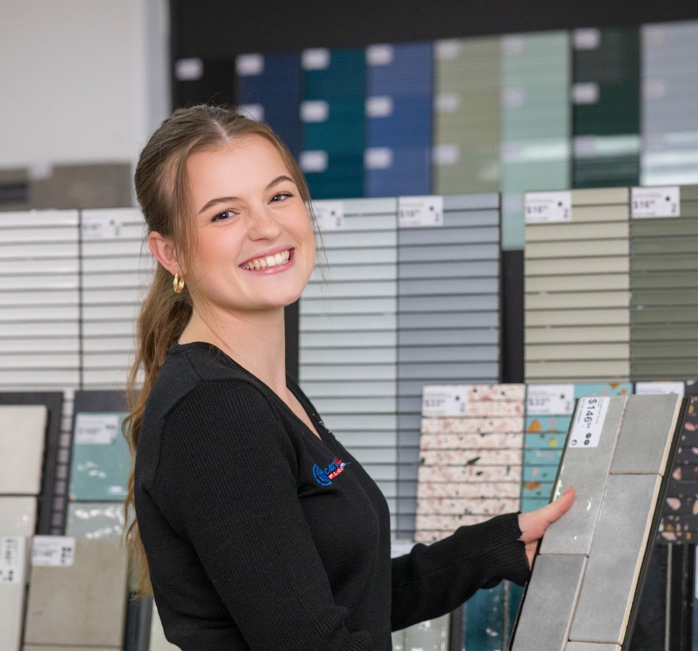 Woman in a tile store smiles, holding up a tile sample. Various tiles are displayed in the background.