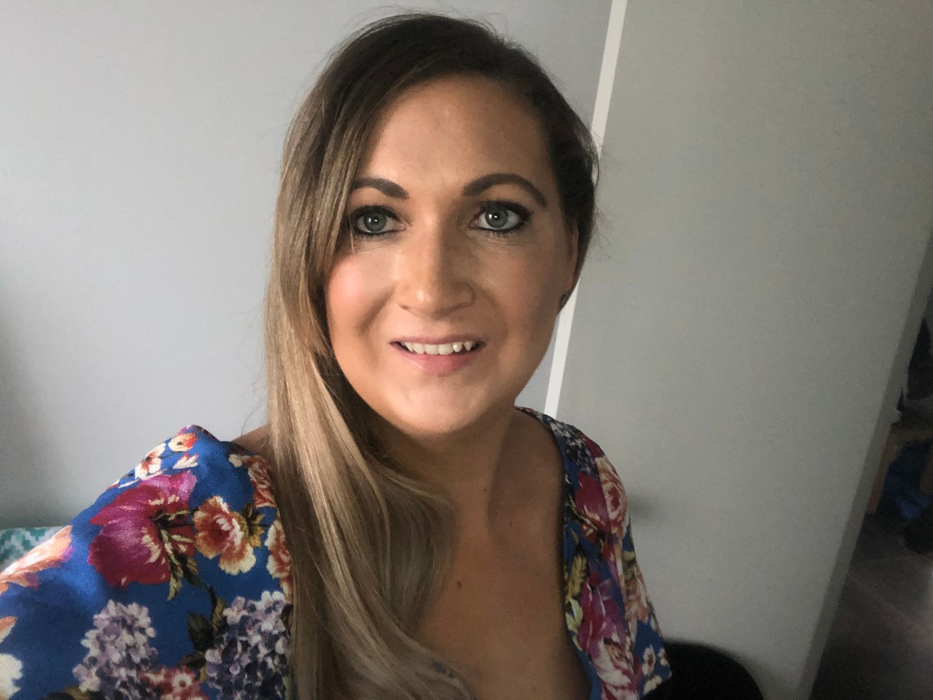Woman with long blonde hair smiles, wearing a blue floral top, against a grey wall.
