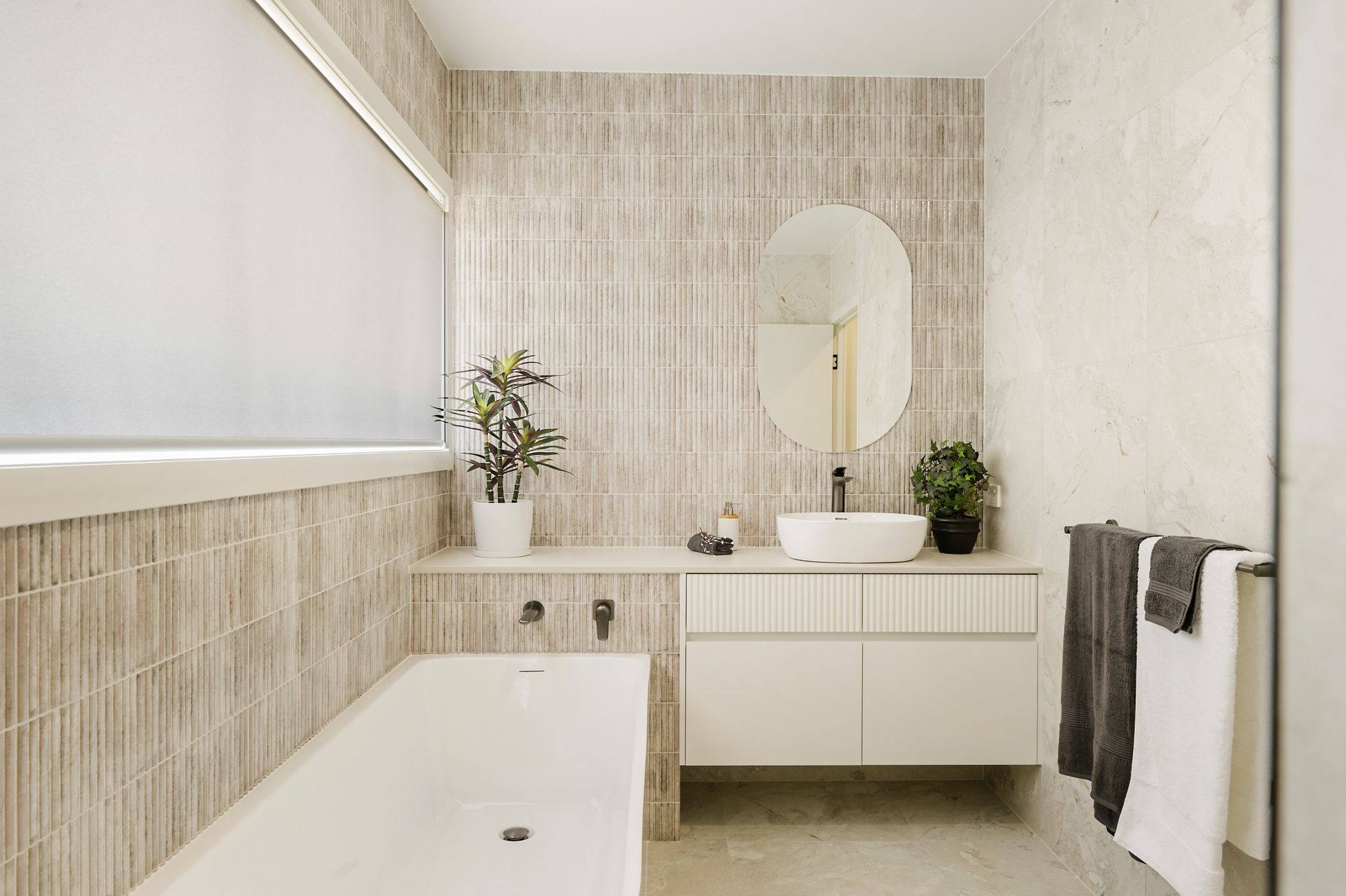 Modern bathroom with neutral tile, white vanity, oval mirror, and bathtub; plants and towels visible.