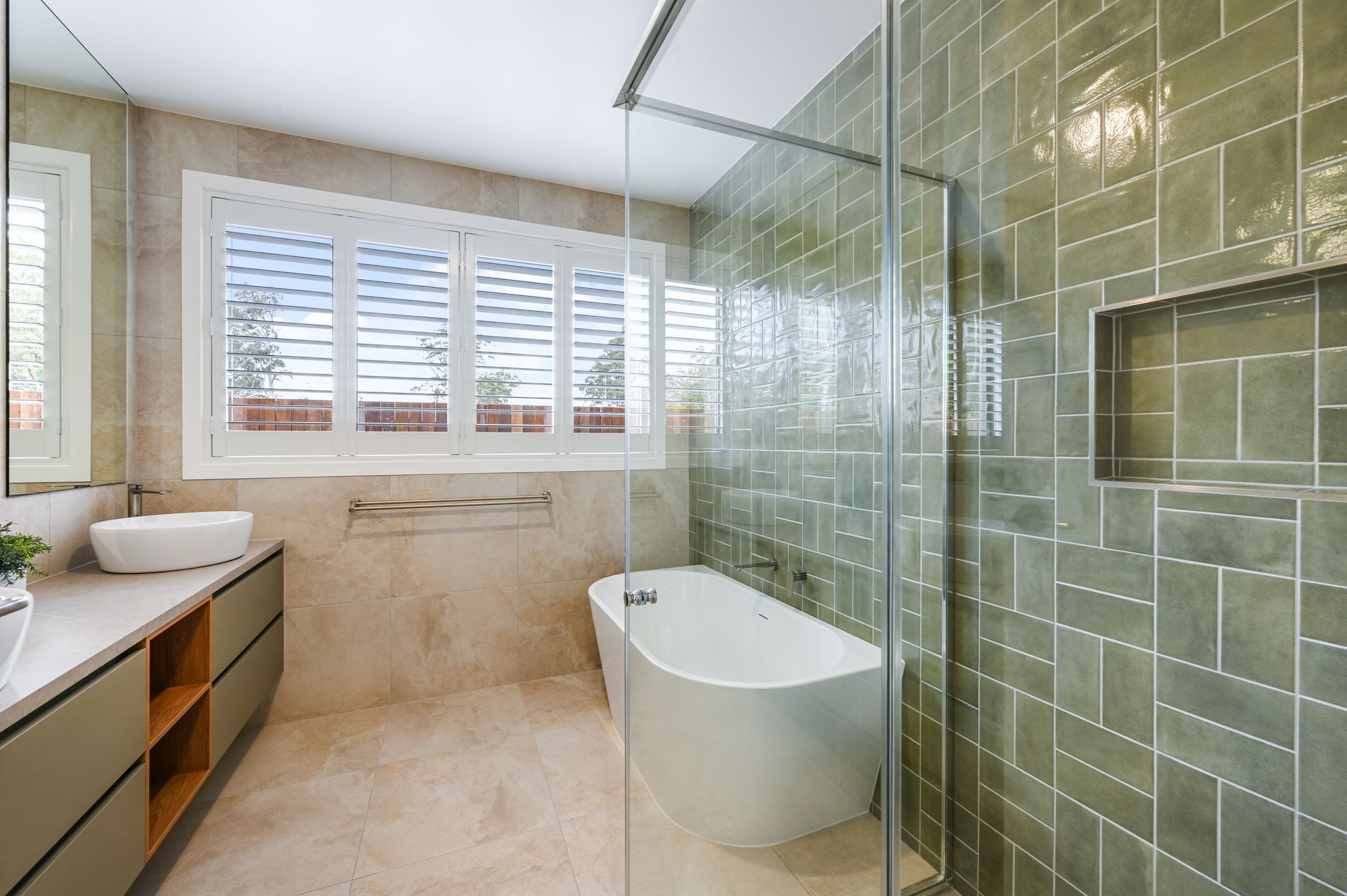 Modern bathroom with green tiled shower, white tub, vanity, and window with shutters.