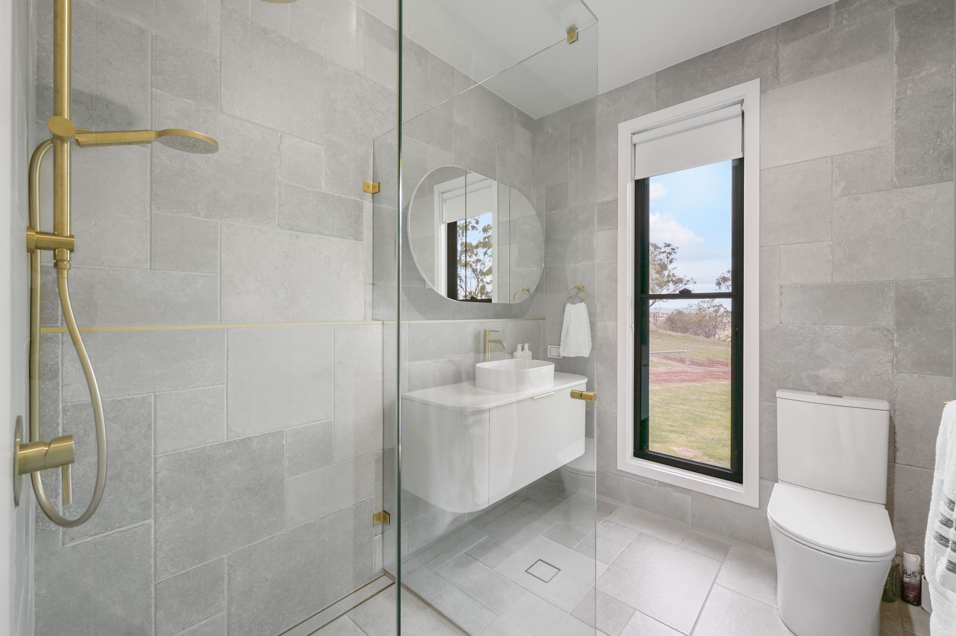 Modern bathroom with grey tiles, gold fixtures, floating vanity, and a window with an outdoor view.