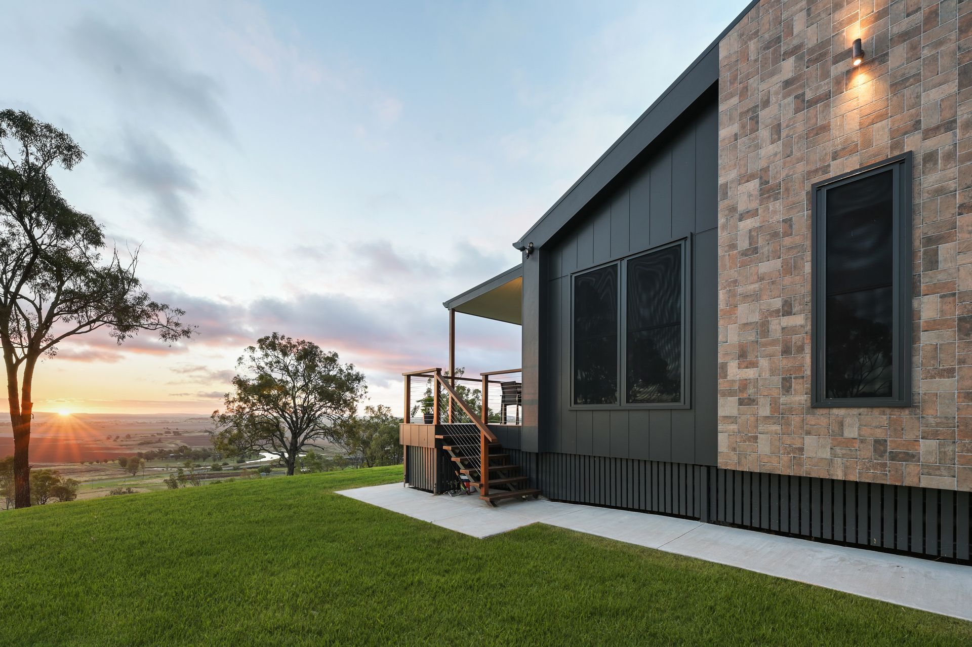 Modern house with stone and dark siding, porch, green lawn, sunset.
