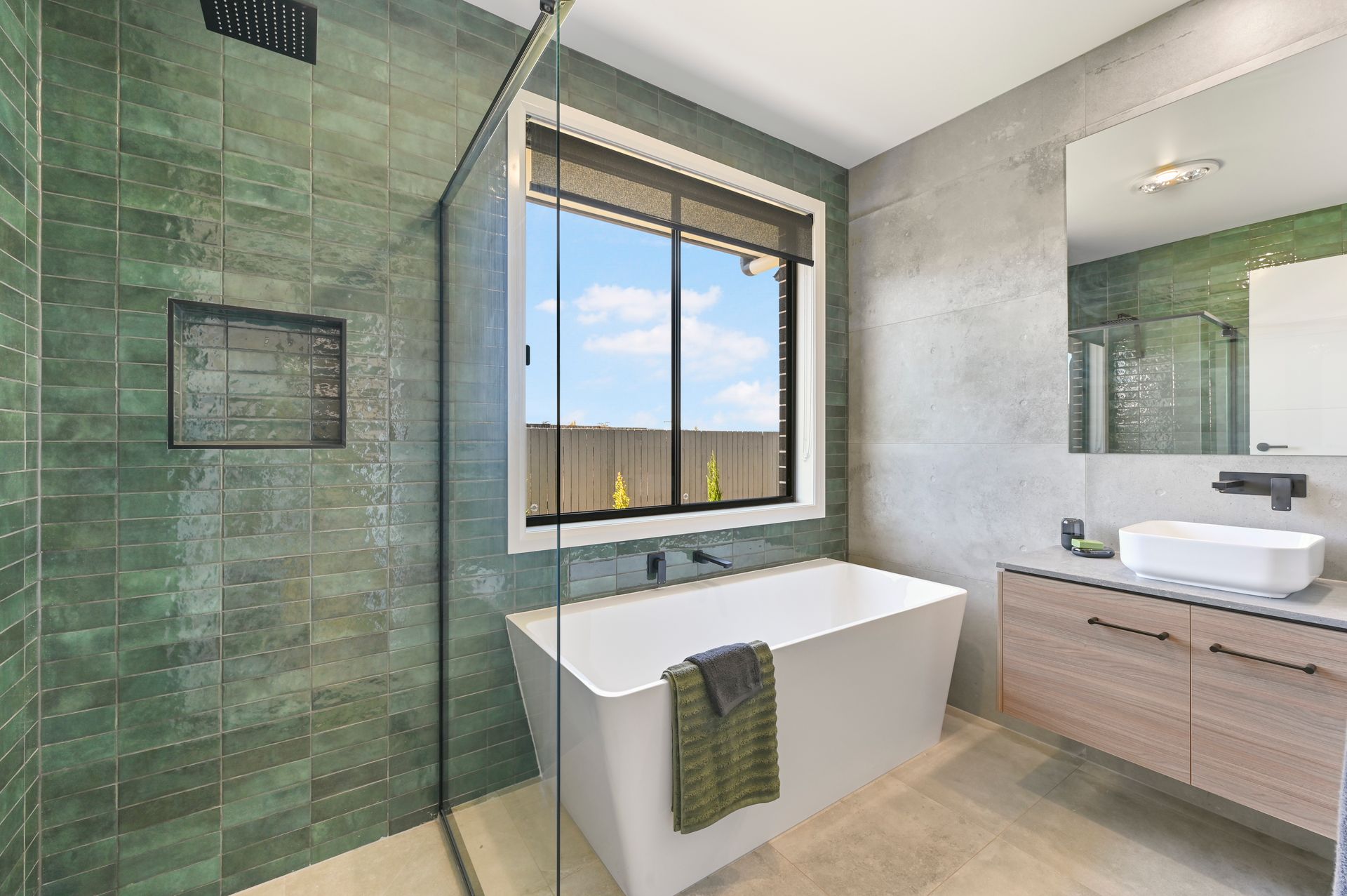 Bathroom with green tile shower, white tub, window with view, and wood vanity.