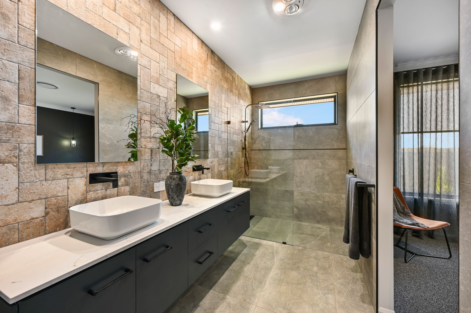 Modern bathroom with stone-like wall tiles, two sinks on a dark cabinet, and a glass shower. Doorway to a room with curtains.