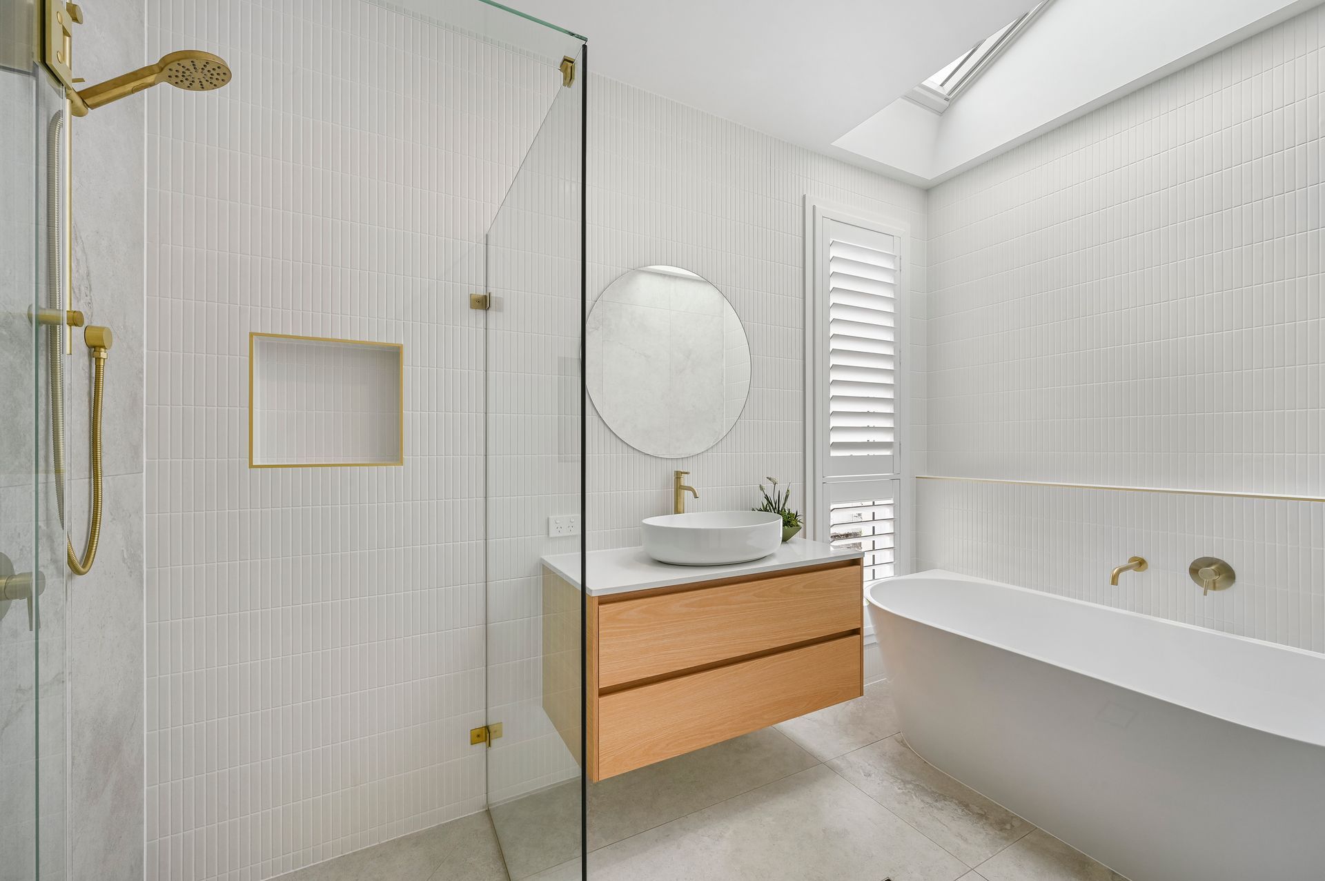 Modern bathroom with white and gold accents: shower, floating vanity, tub, skylight, and shuttered window.
