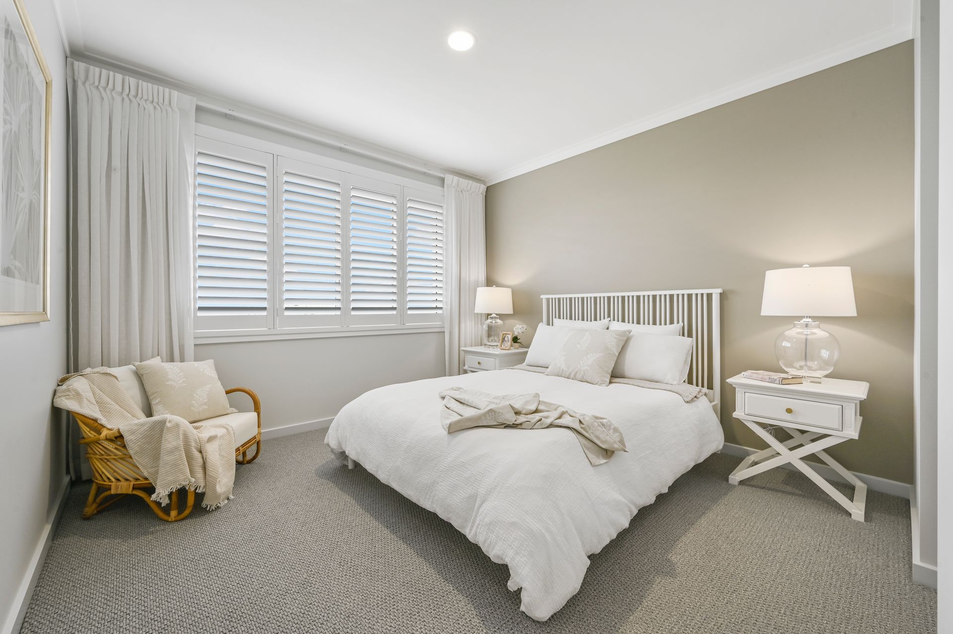 Bedroom with white bedding, a rattan chair, and window shutters. Grey carpet and walls.