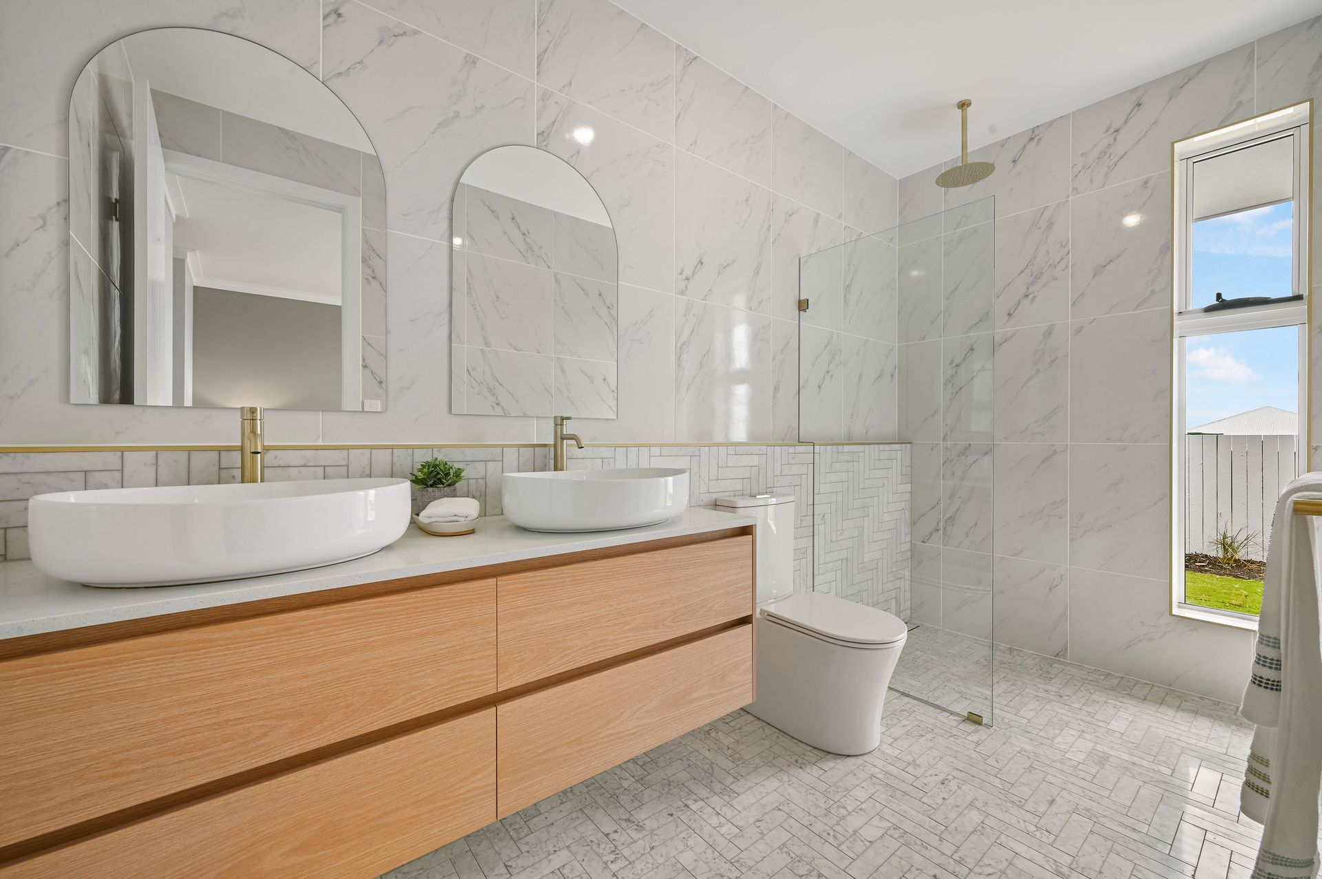 Modern bathroom with marble walls, wood vanity, and dual vessel sinks. Two arched mirrors and a glass shower.