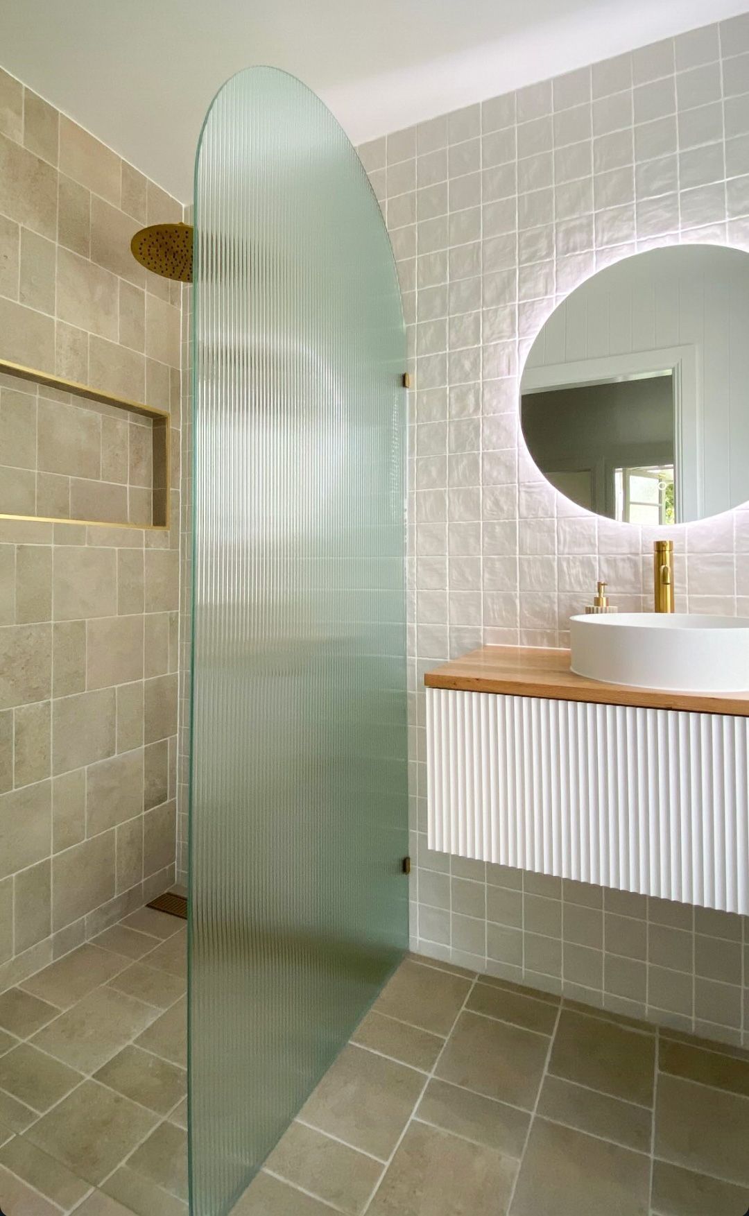Bathroom with fluted glass shower door, gold fixtures, and a floating vanity with a round mirror.