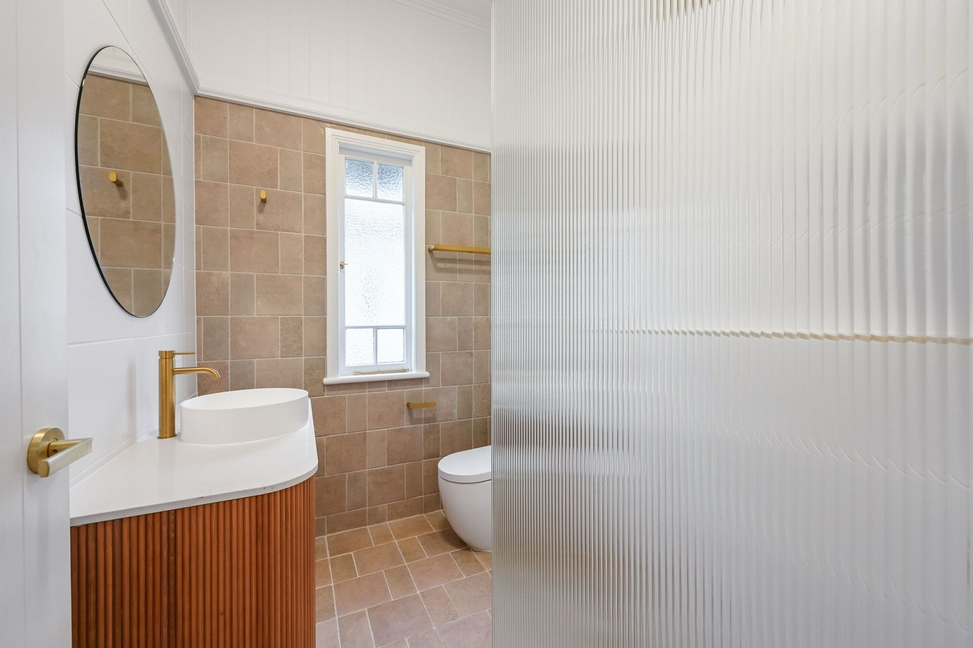 Bathroom with tan tiles, a gold faucet and fixtures, and a ribbed glass shower wall.