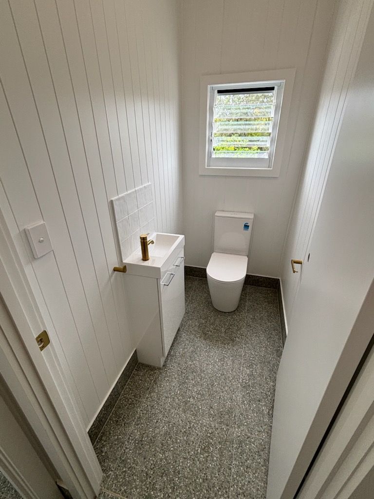 Small, modern bathroom with white walls and fixtures. Toilet and sink on speckled floor, window above toilet.