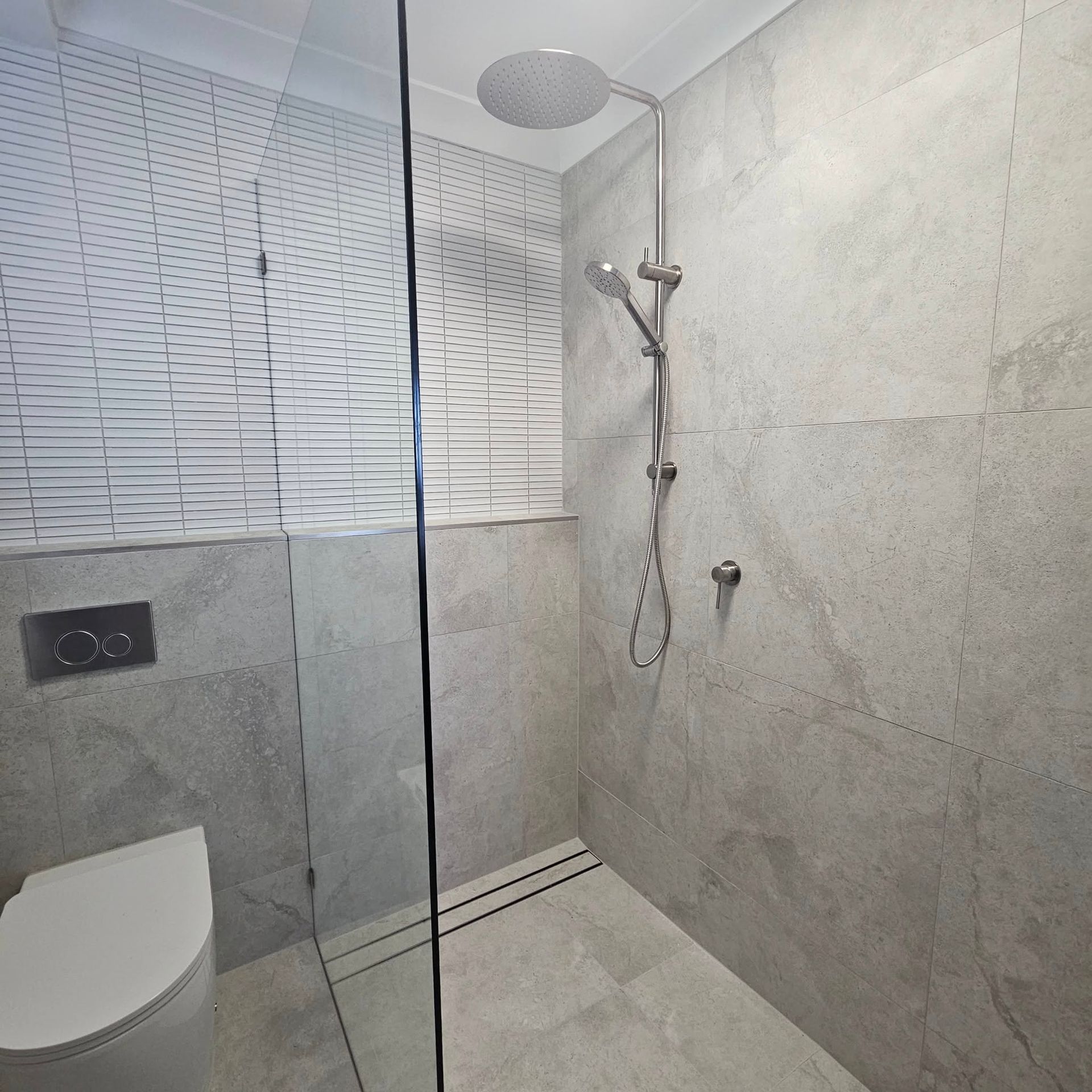 Modern shower with grey tiled walls and a glass door, next to a toilet.