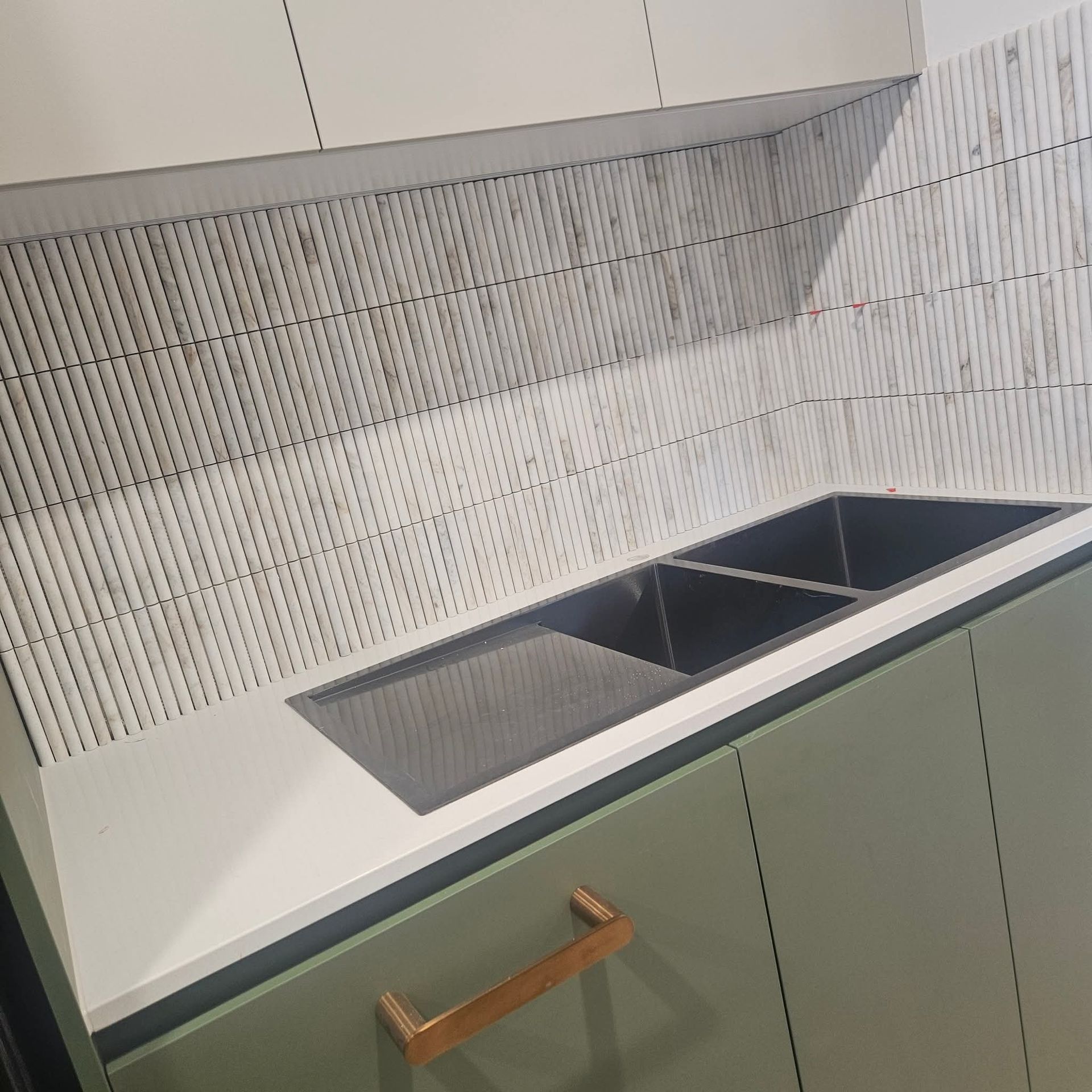 A kitchen with a green cabinet, white countertop, and a black sink. The backsplash is a textured tile.