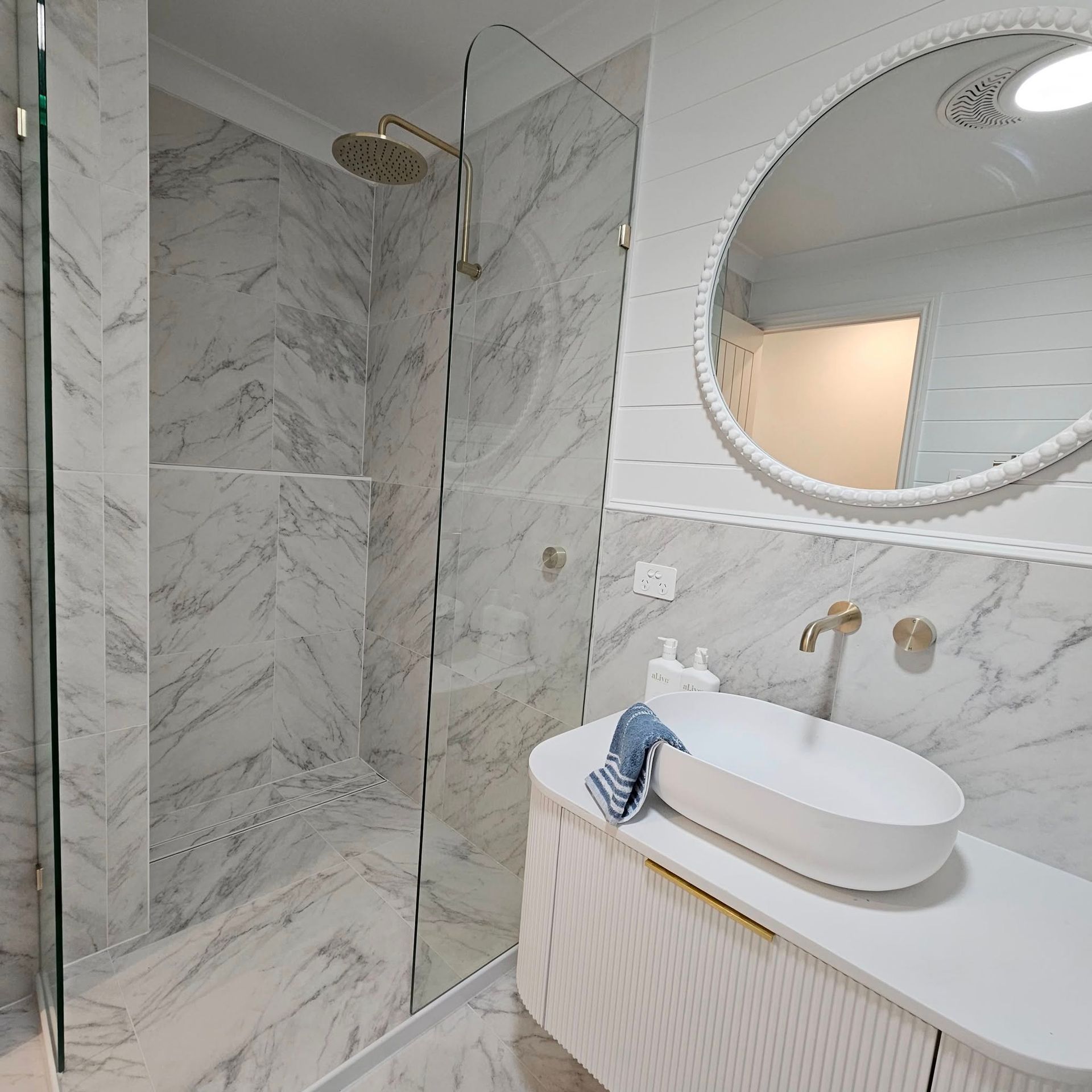Modern bathroom with white marble tiles, glass shower, white vanity, and round mirror.