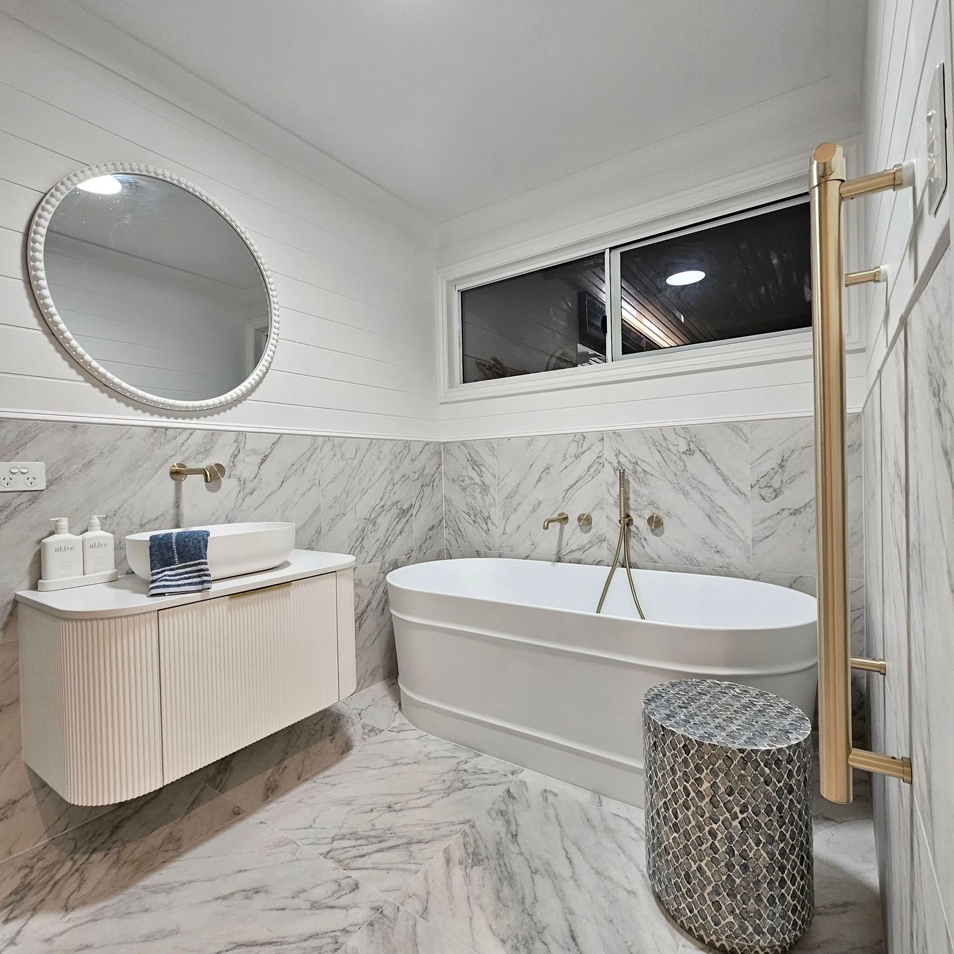 Luxurious white marble bathroom with a soaking tub, vanity, round mirror, and gold fixtures.