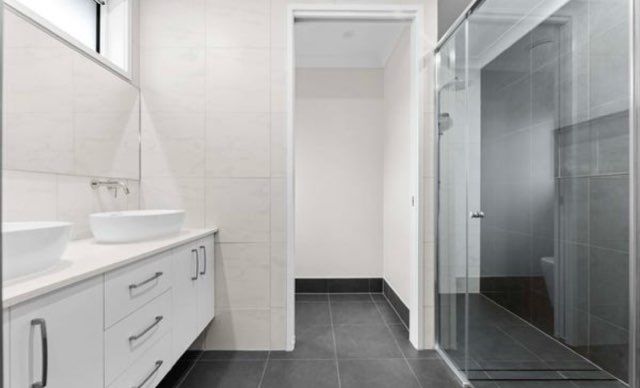 Modern bathroom: white vanity with two sinks, grey tiled floor, glass shower, doorway to a toilet.