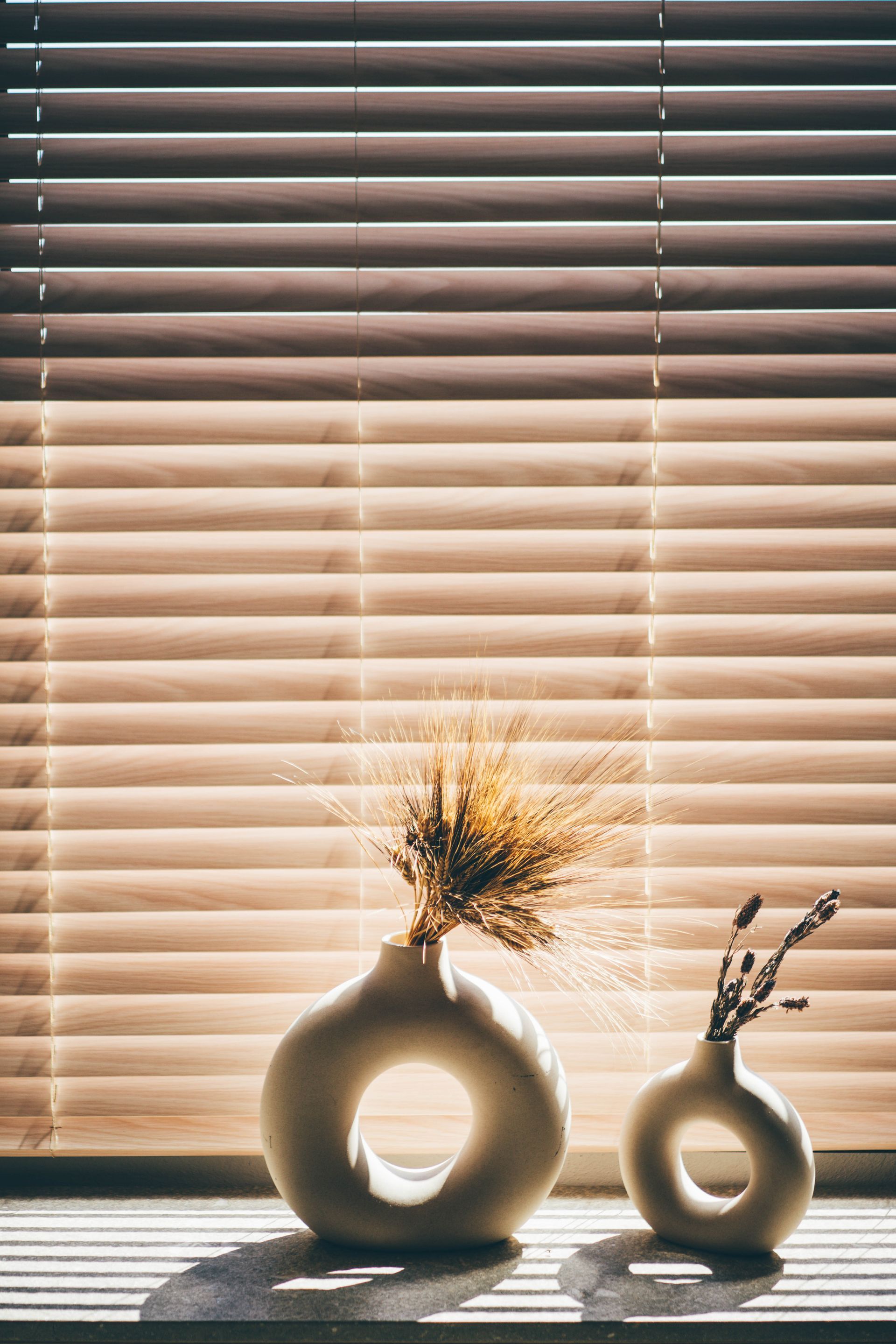 Two white, donut-shaped vases with dried plants on a windowsill with slatted blinds, sunlight.