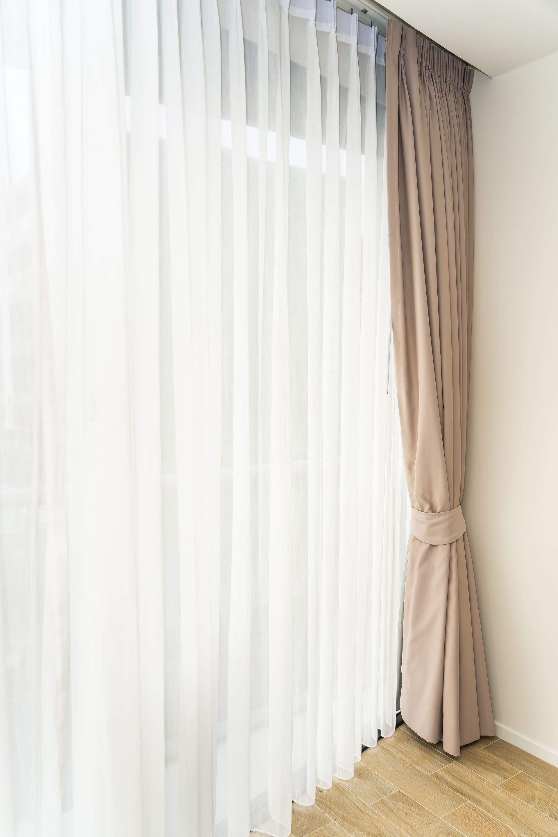 Beige and white curtains framing a window, brown curtain tied back, wood floor.