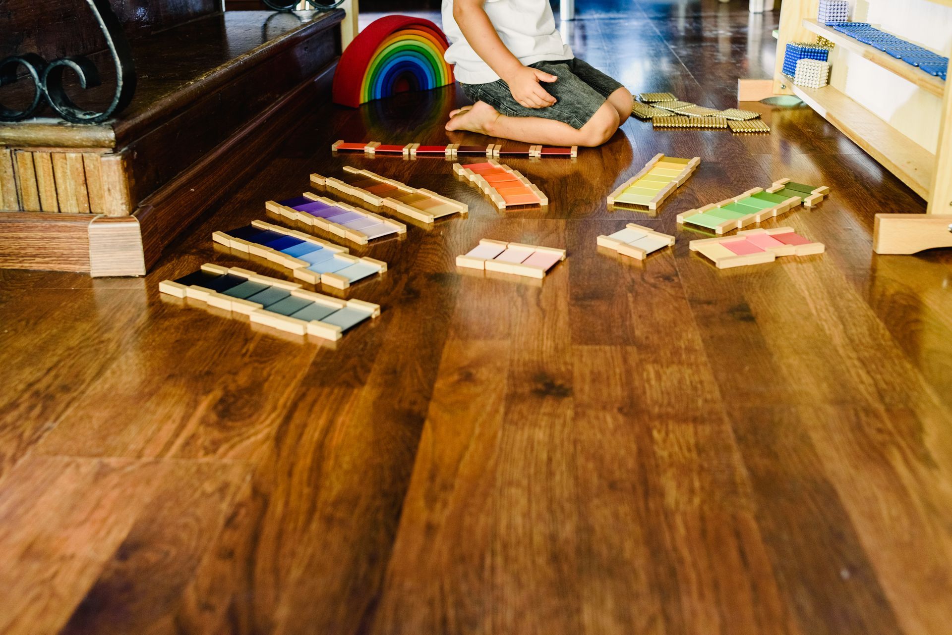 A child playing with coloured wooden blocks on a hardwood floor, a rainbow-shaped toy is visible in the background.