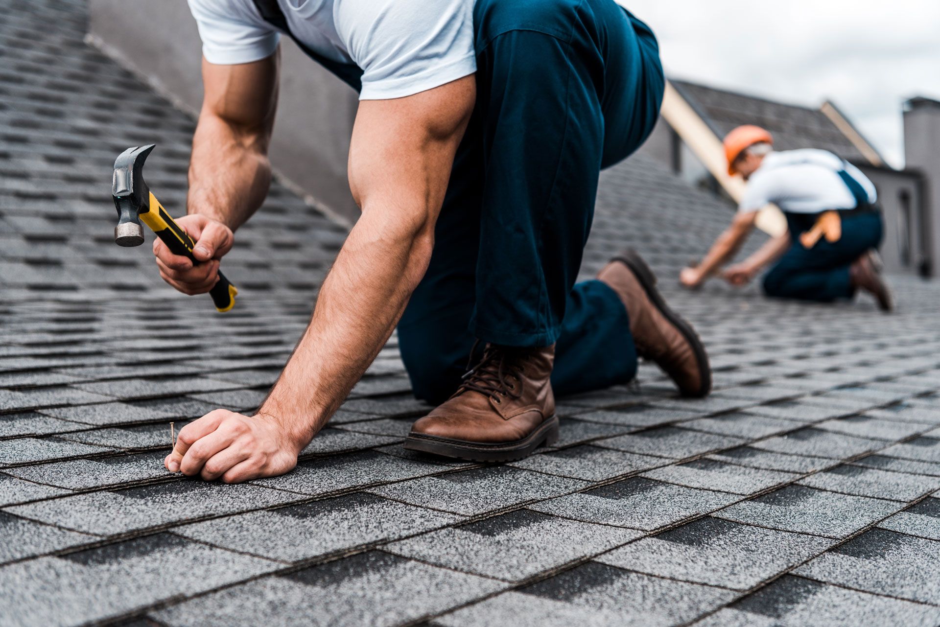 Two roofers kneeling on a roof, one hammering shingles.
