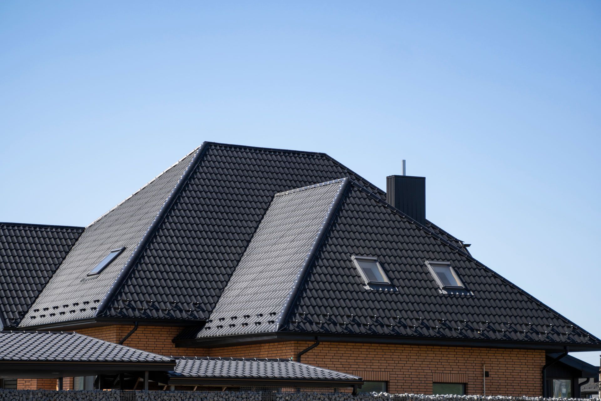 Dark gray tiled roof of a house with two skylights and a chimney against a blue sky.