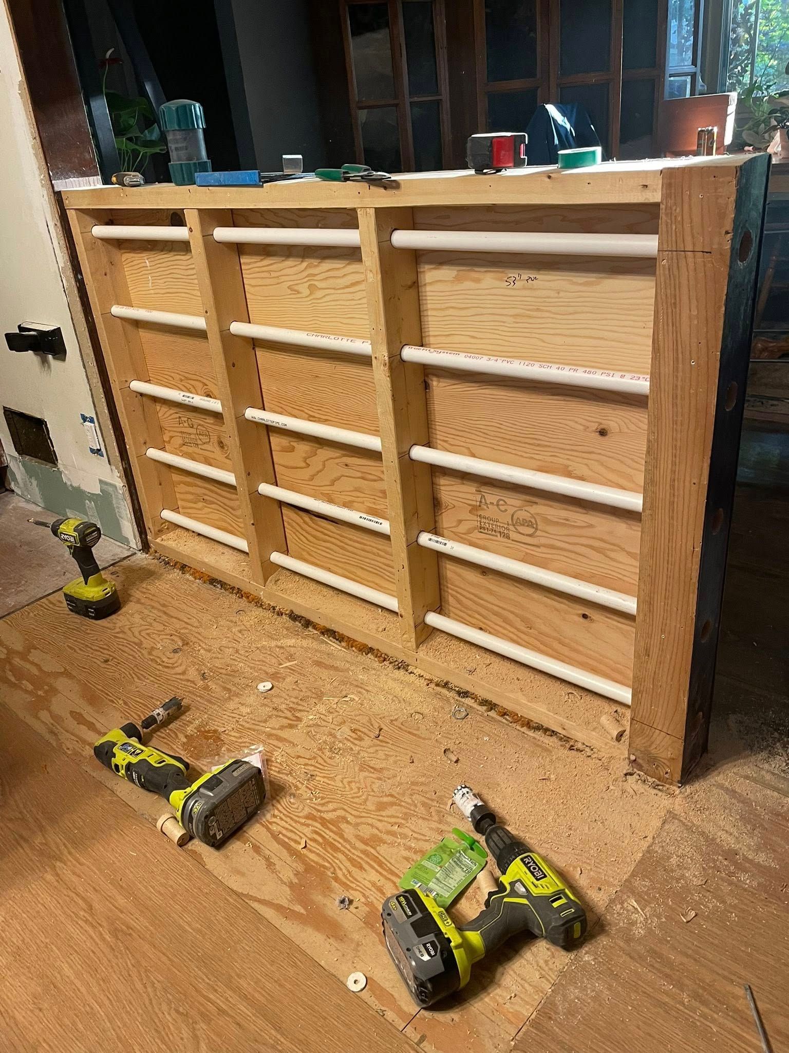 Wooden structure with horizontal white pipes, surrounded by tools and wood shavings in a construction setting.