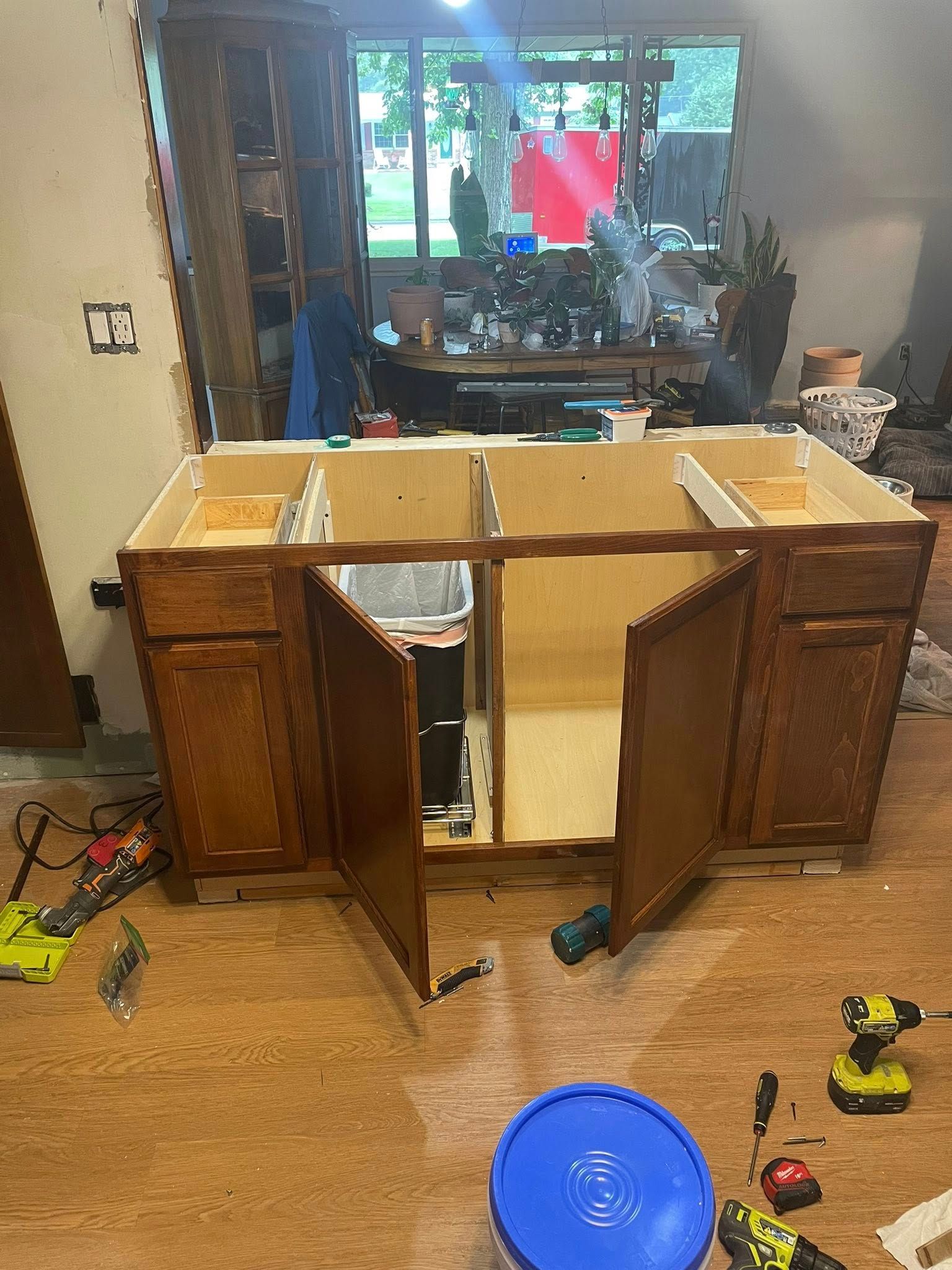 Kitchen cabinet under construction, brown wood, with open doors, trash bin inside. Tools and blue bucket on the floor.