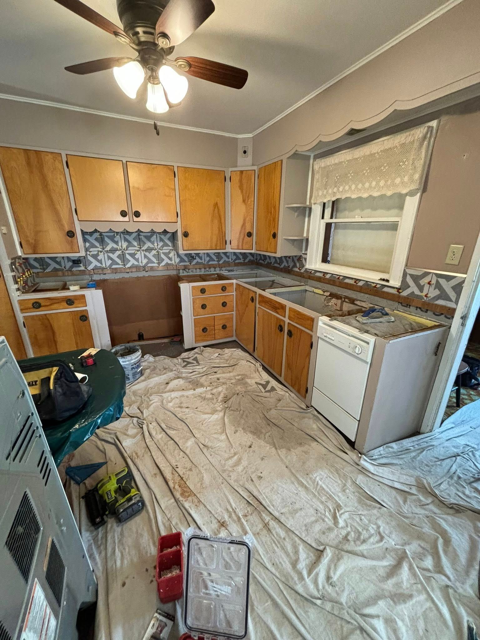 Kitchen under renovation, featuring wooden cabinets, a white dishwasher, and paint tarps on the floor.