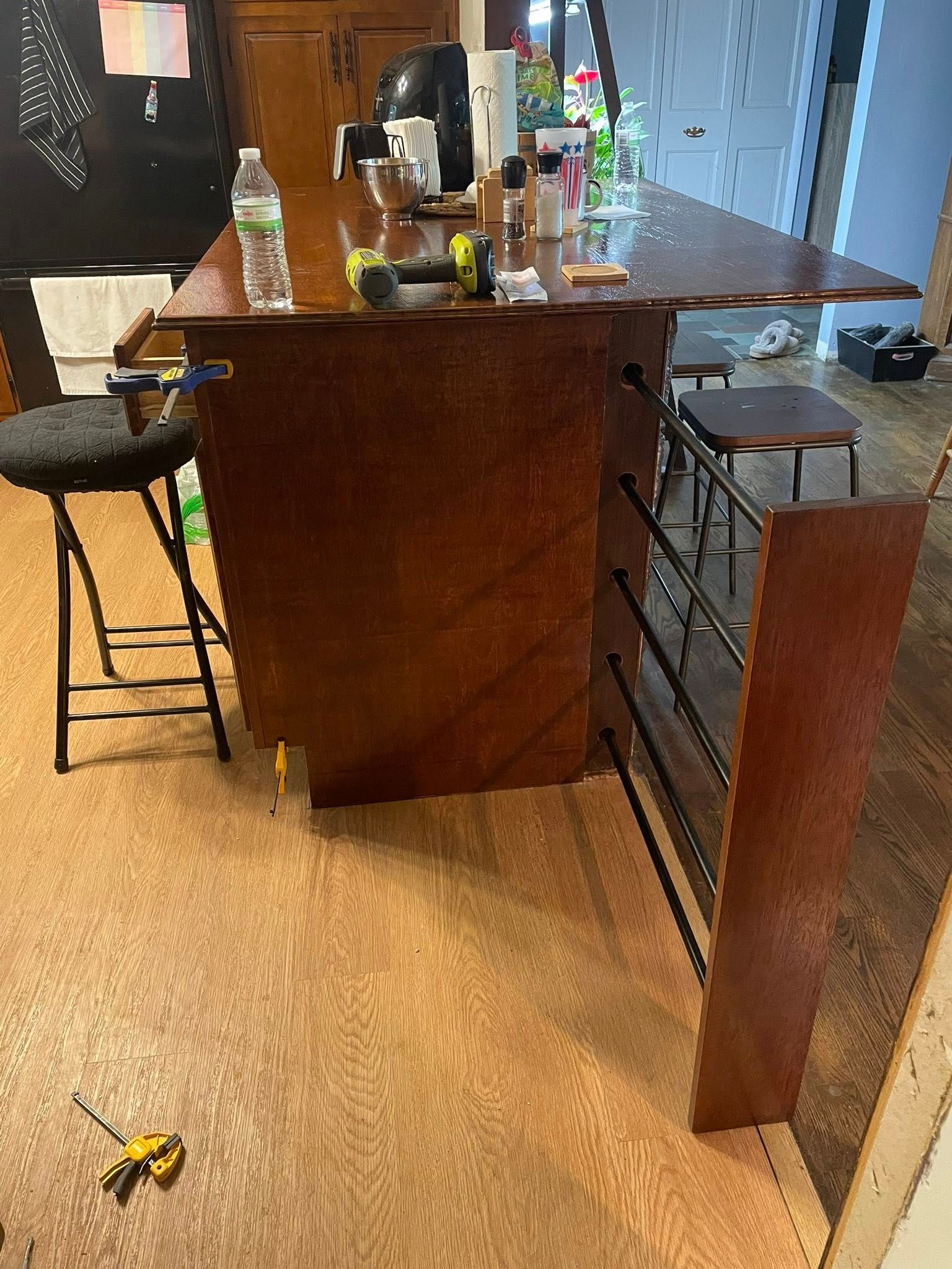 A wooden bar with stools and a built-in footrest. Tools and bottles sit on the bar.
