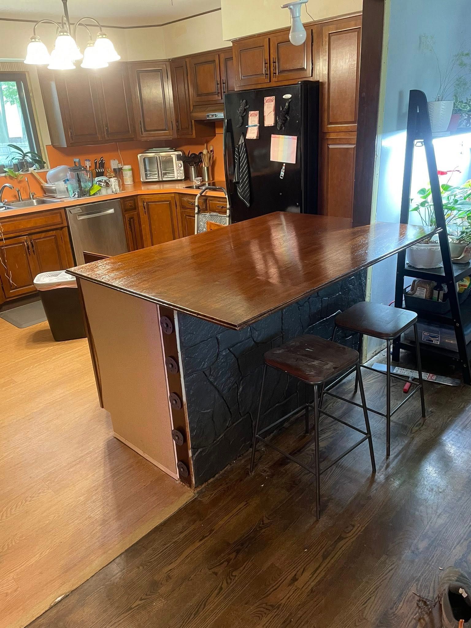 Kitchen with brown cabinets, island, and stool. Refrigerator, dishwasher visible. Brown floors.