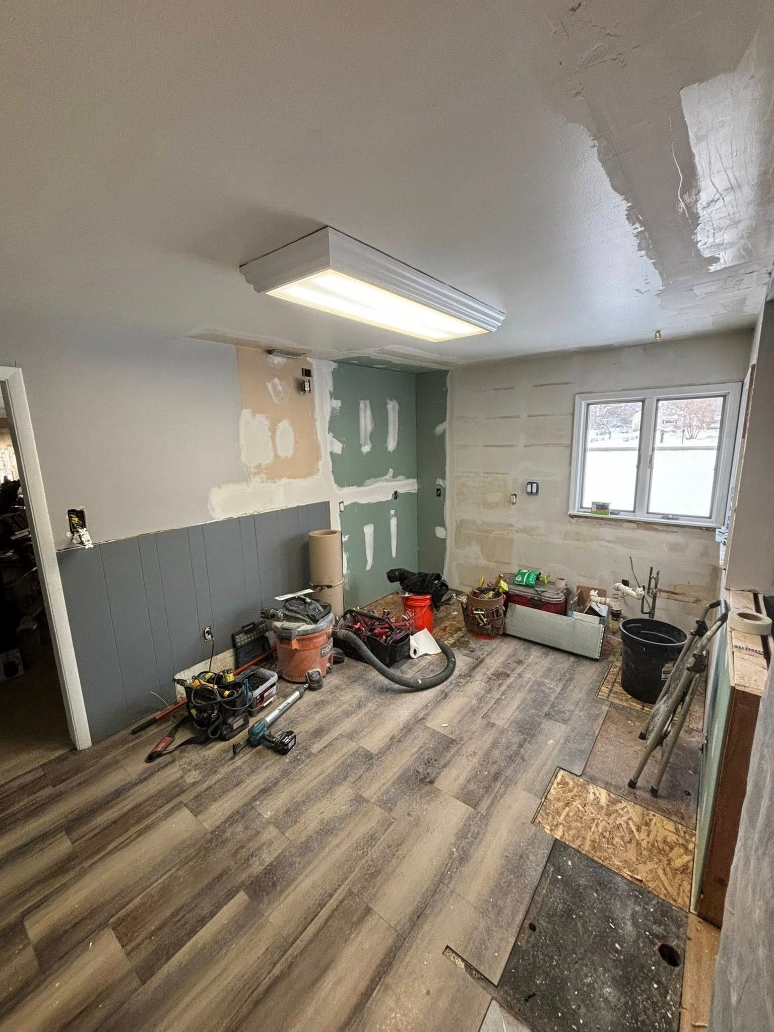Room under construction with exposed walls, tools, and new flooring. Fluorescent light on ceiling.