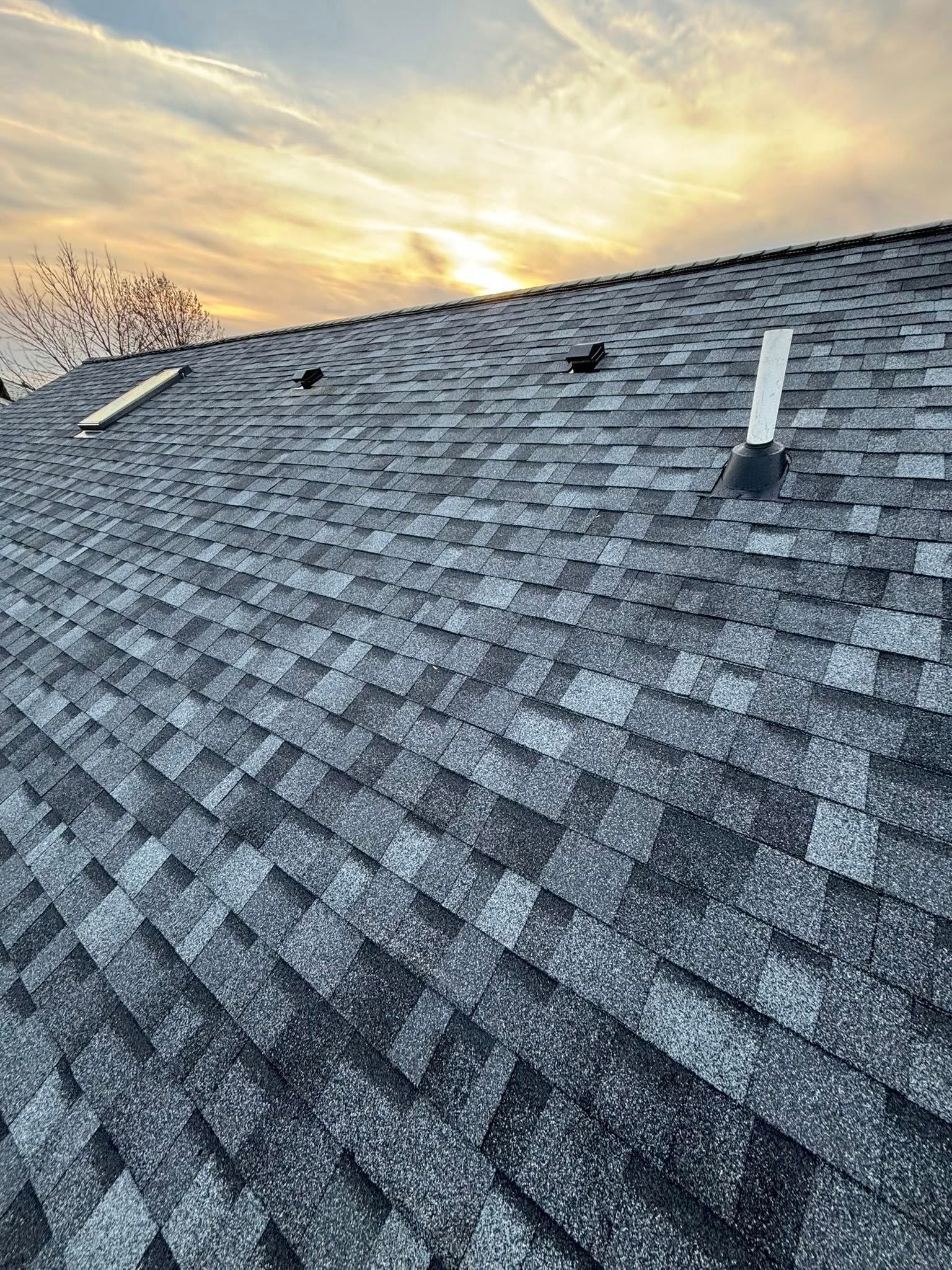 Asphalt shingle roof with vents against a sunset sky.